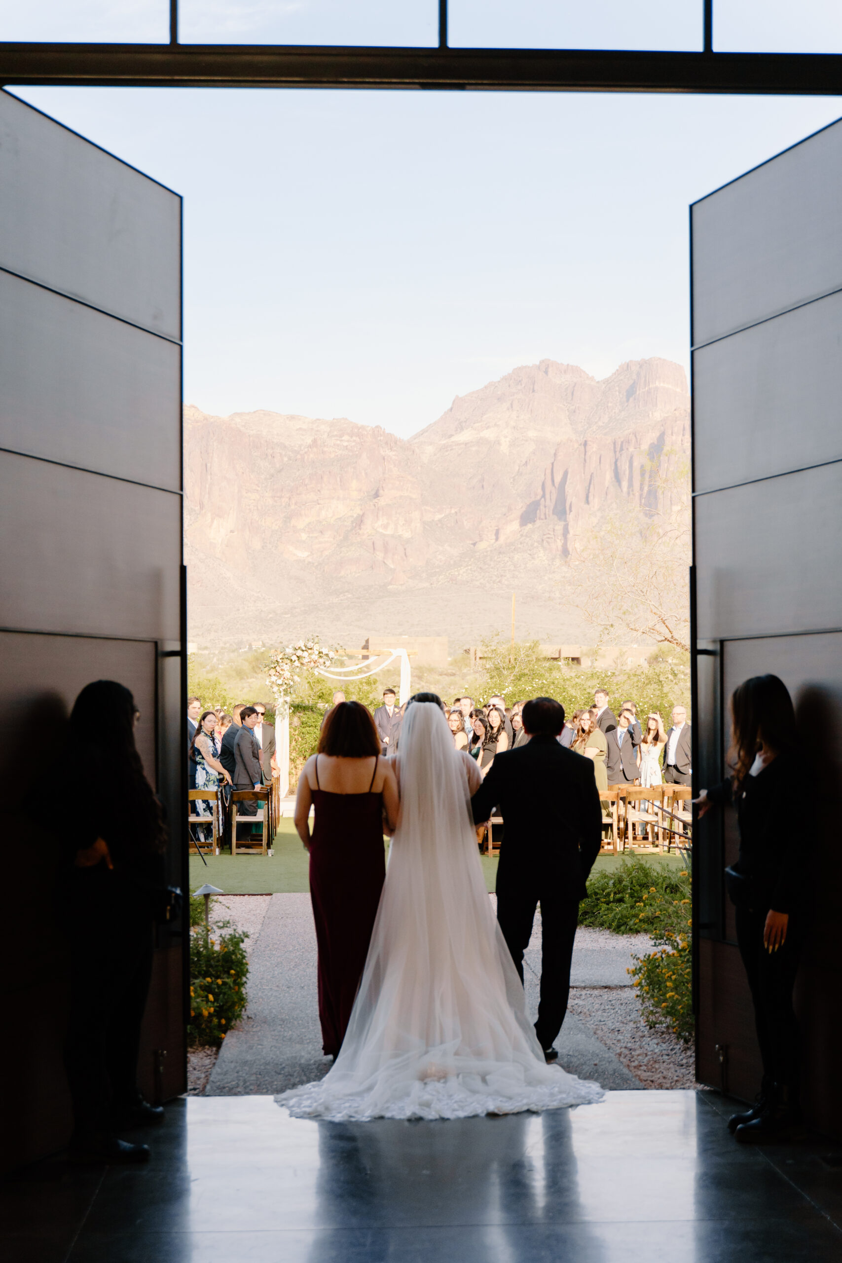 The bride with her parents by her side during her ceremony at The Paseo, Arizona, framed by large open doors with mountain scenery ahead.