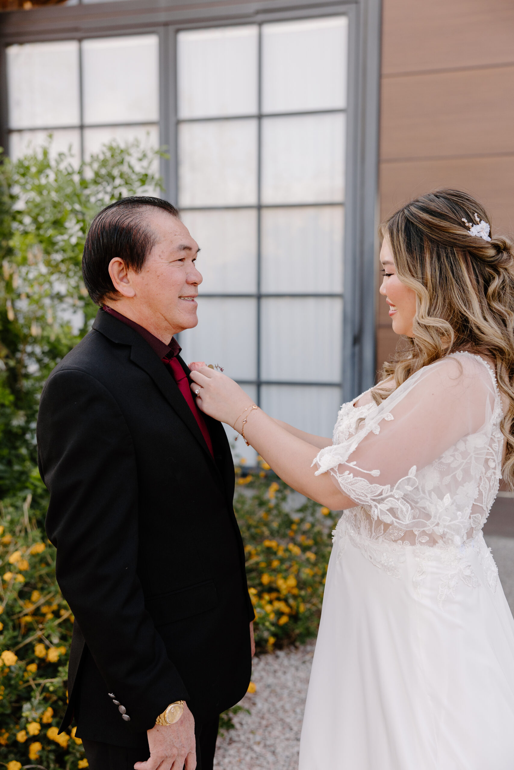 A bride in a white lace dress adjusts the tie of a man in a black suit and red shirt. They stand outdoors, smiling at each other warmly.