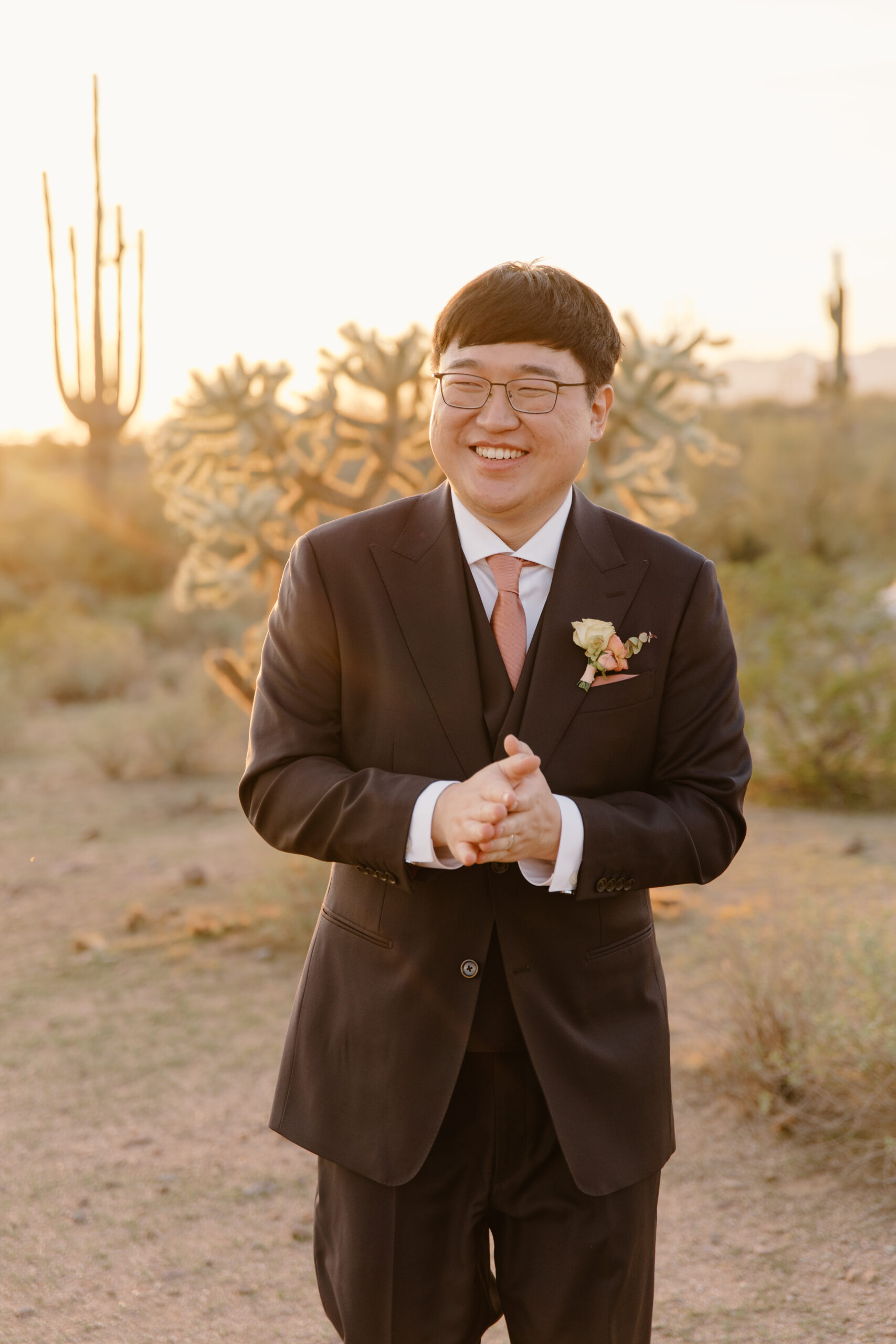 The groom smiling and clapping his hands together during a golden-hour portrait session at The Paseo, Arizona, desert plants softly surrounding him.
