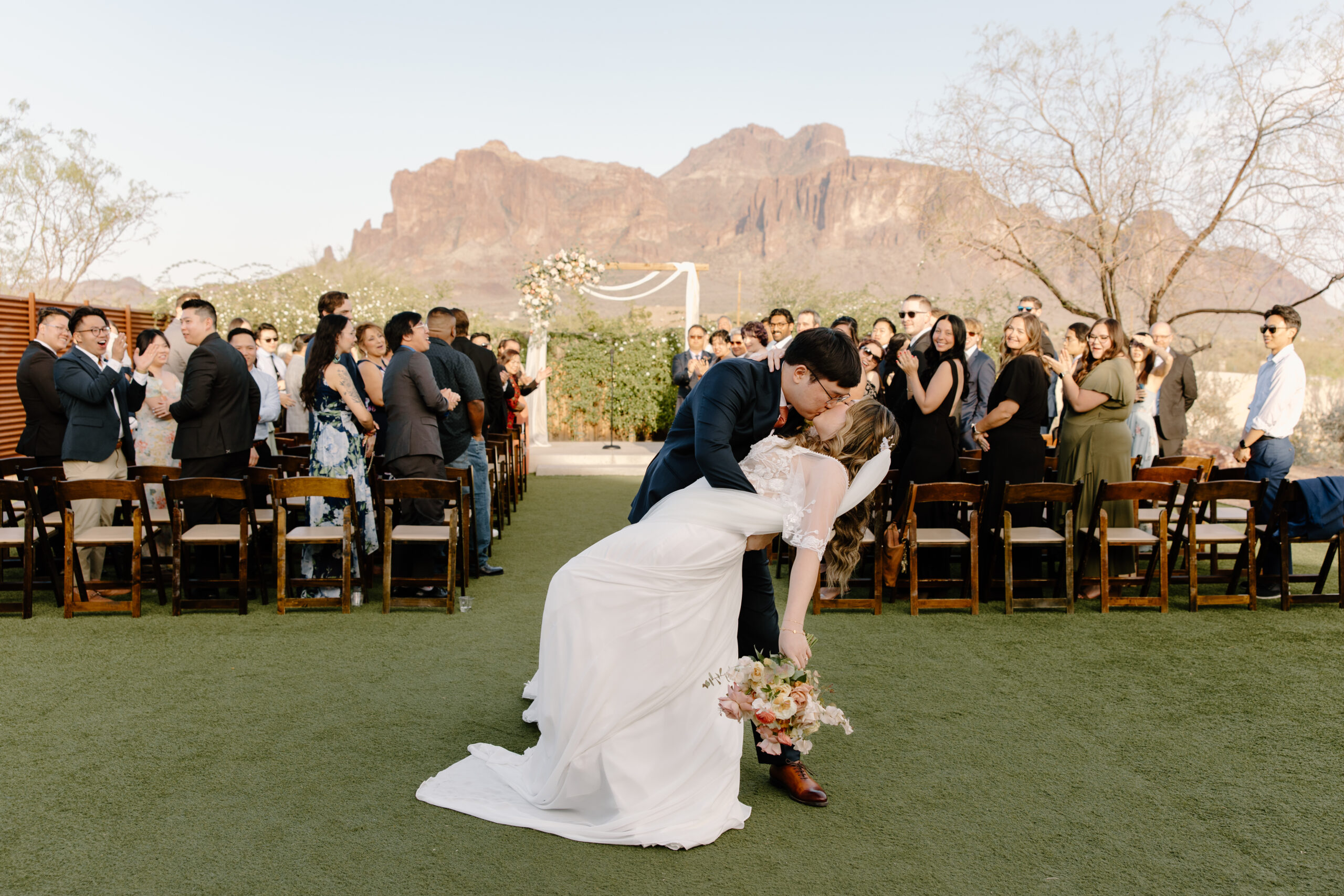 The groom leaning the bride into a romantic dip kiss at their outdoor wedding ceremony at The Paseo, Arizona, wooden chairs lining the turf aisle.