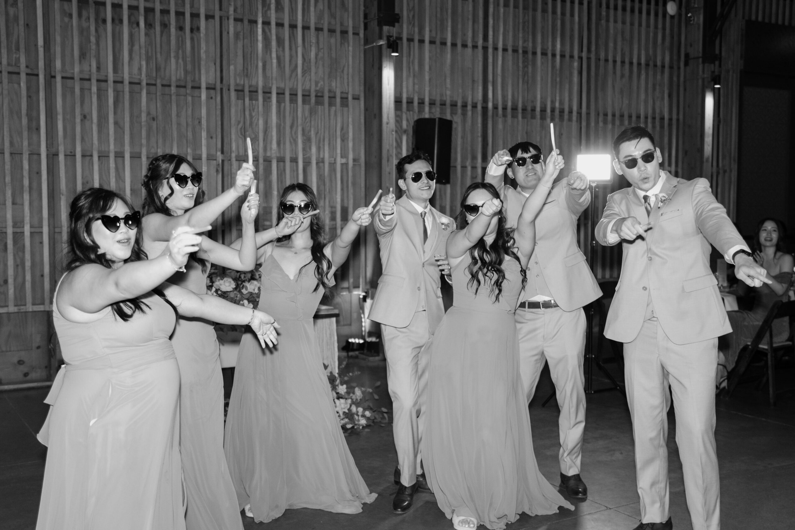 A group of seven joyful people wearing sunglasses strike playful poses at a wedding reception. They are dressed in formal suits and gowns.