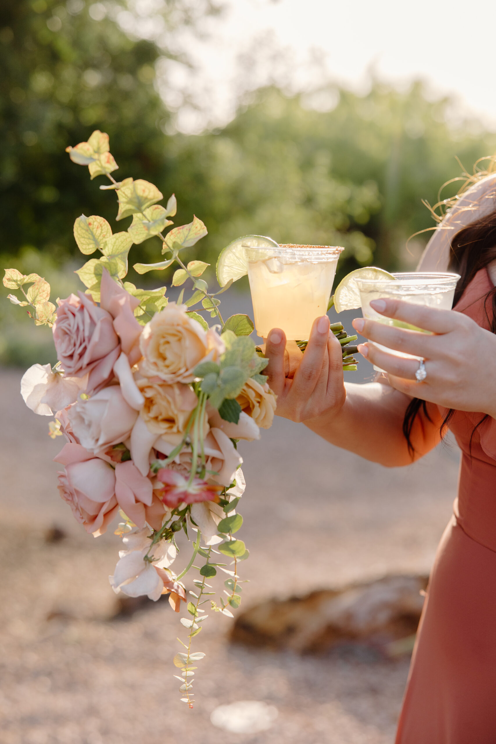 A person in a peach dress holds a bouquet of pink roses and greenery, with two cocktail glasses garnished with lime. Warm, sunny outdoor setting at The Paseo, Arizona.