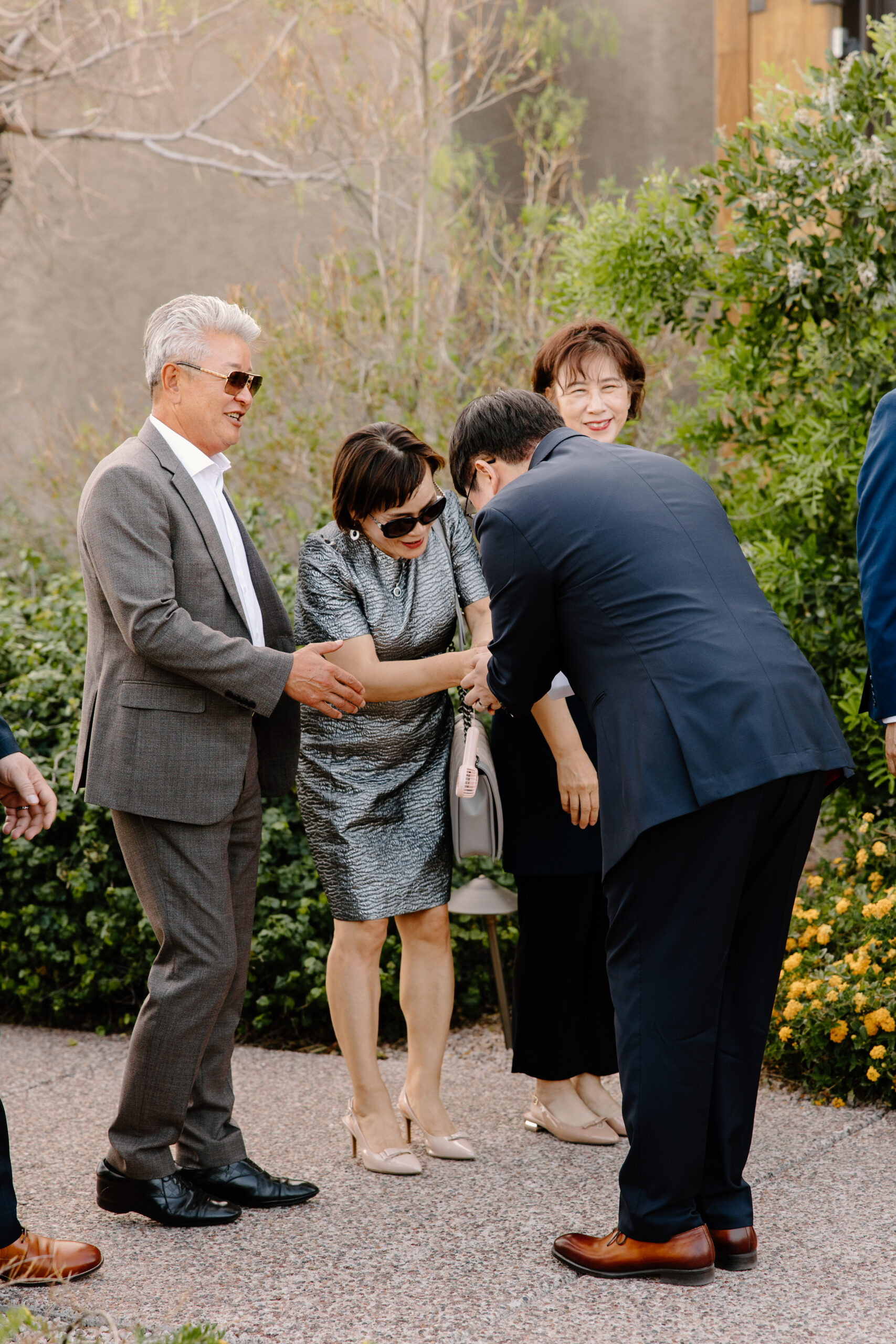 A group of four people outdoors. One person in a suit bows while shaking hands with a smiling woman in sunglasses. Others smile, surrounded by greenery.