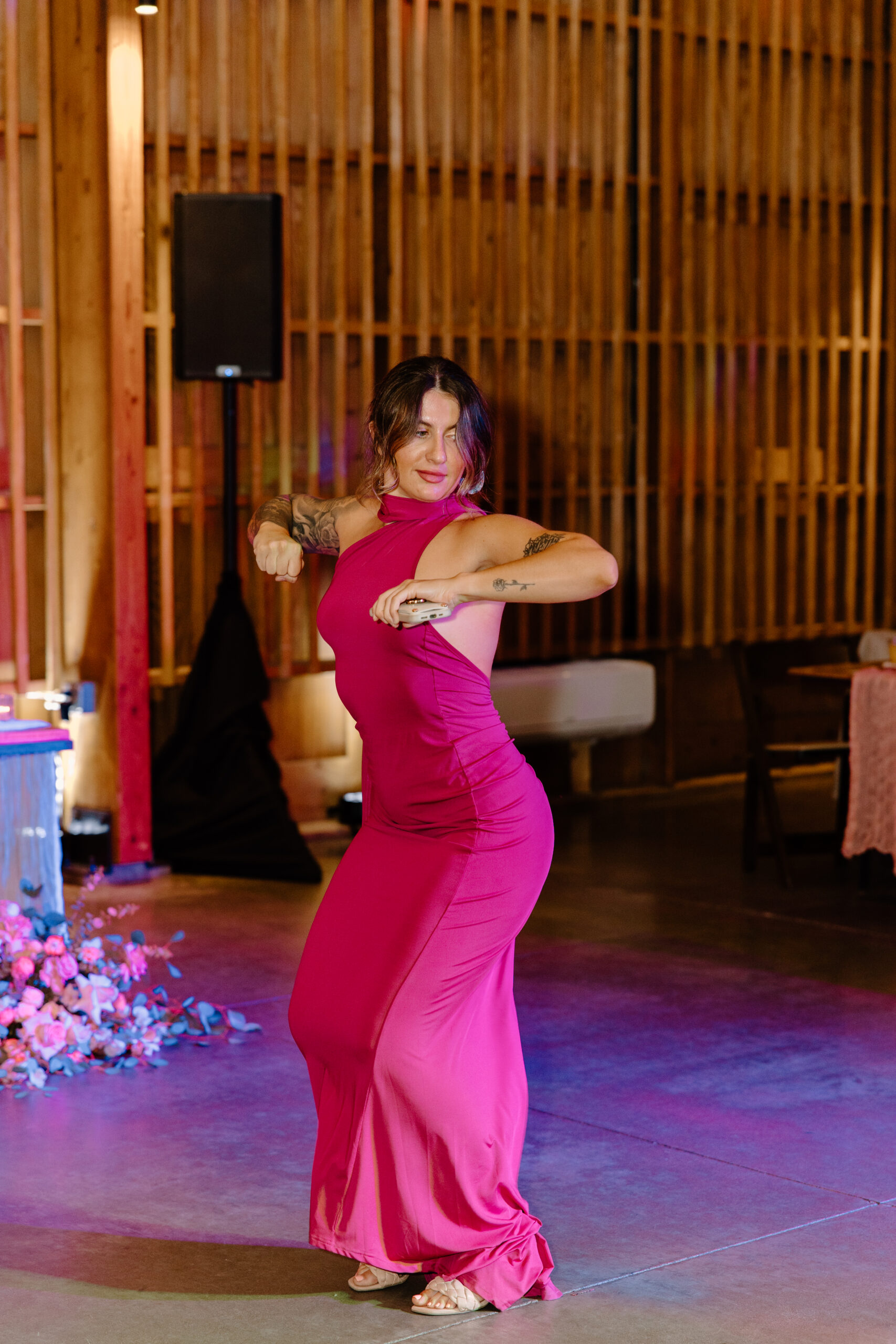 A woman in a vibrant pink dress dances energetically indoors. The setting has warm lighting and wooden walls, creating a festive, lively atmosphere.