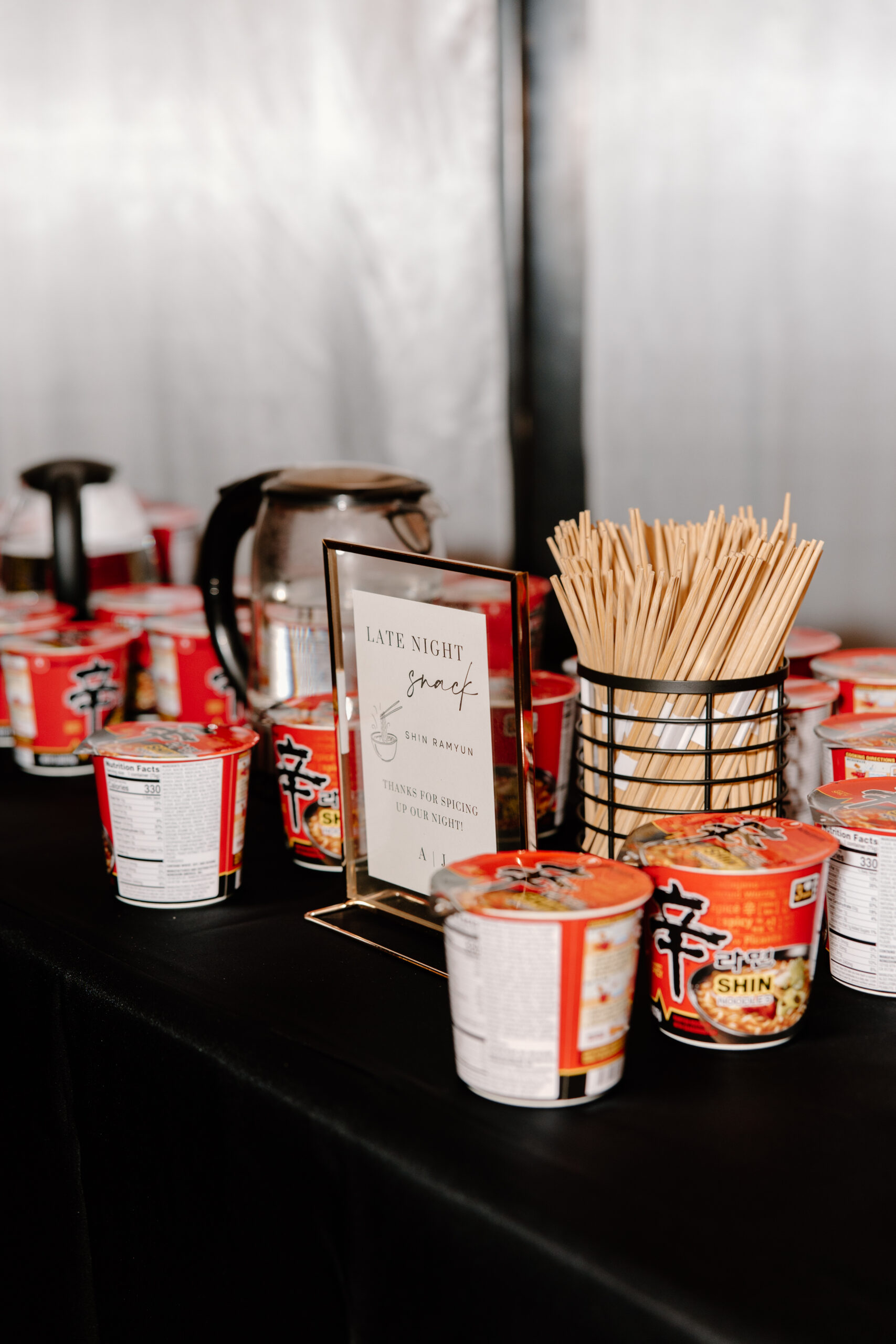 A table with red Shin Ramyun noodle cups, a pot, and chopsticks in a holder. A sign reads "Late Night Snack" offering a cozy atmosphere.