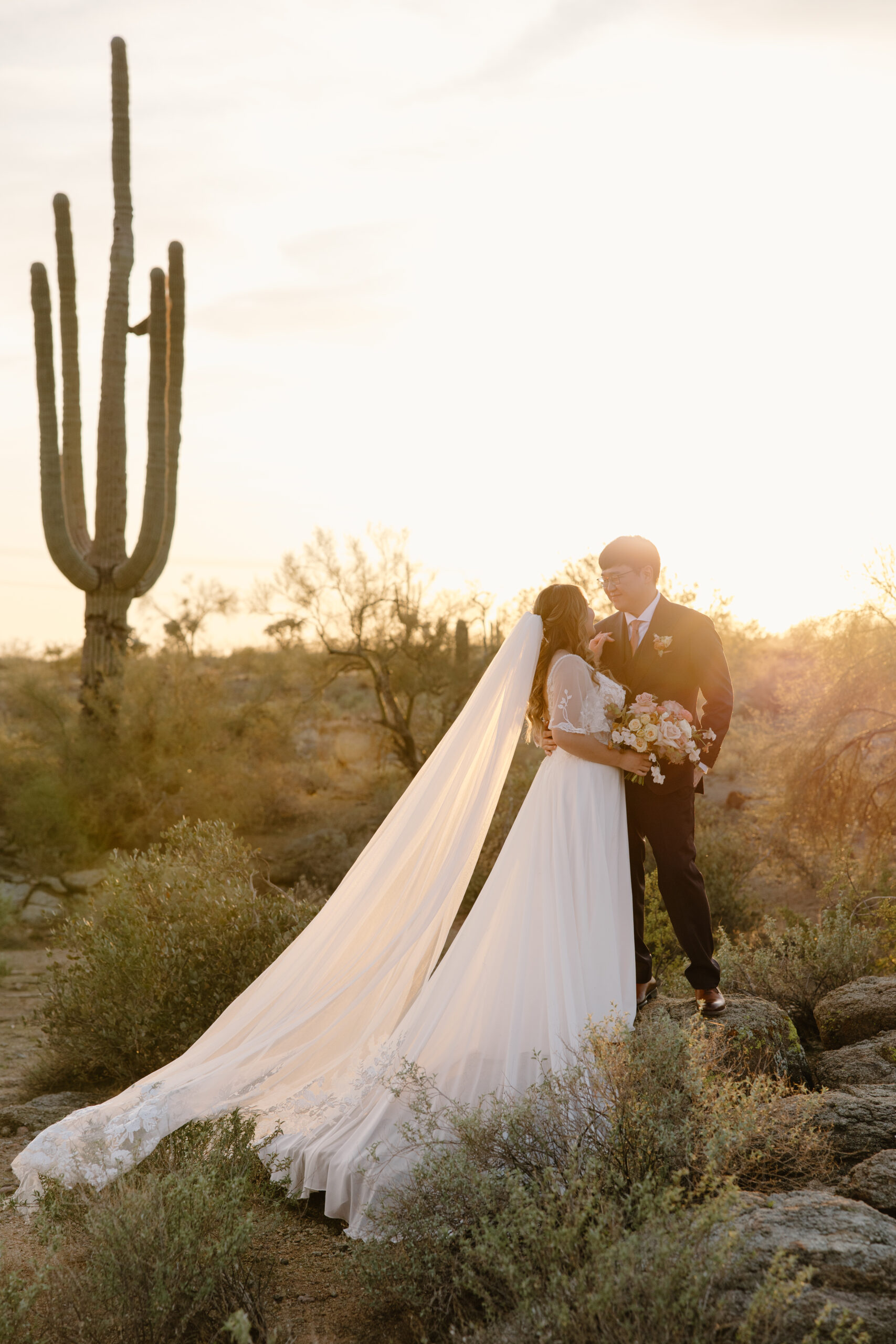 A bride and groom stand together in the Arizona desert at sunset, her long veil flowing as a large cactus rises behind them.