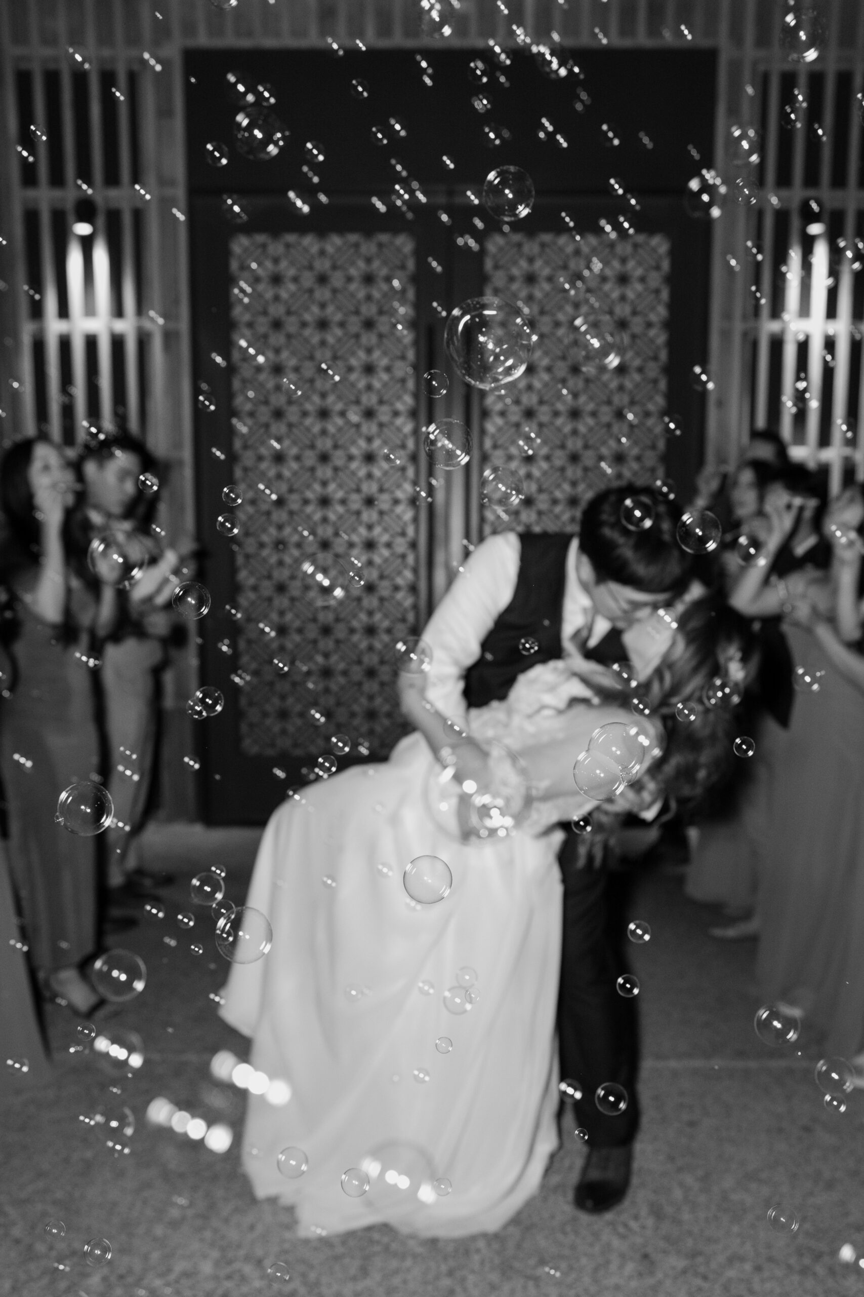 A black-and-white image of the bride and groom sharing a deep, joyful dip kiss while bubbles float around them in front of patterned venue doors.