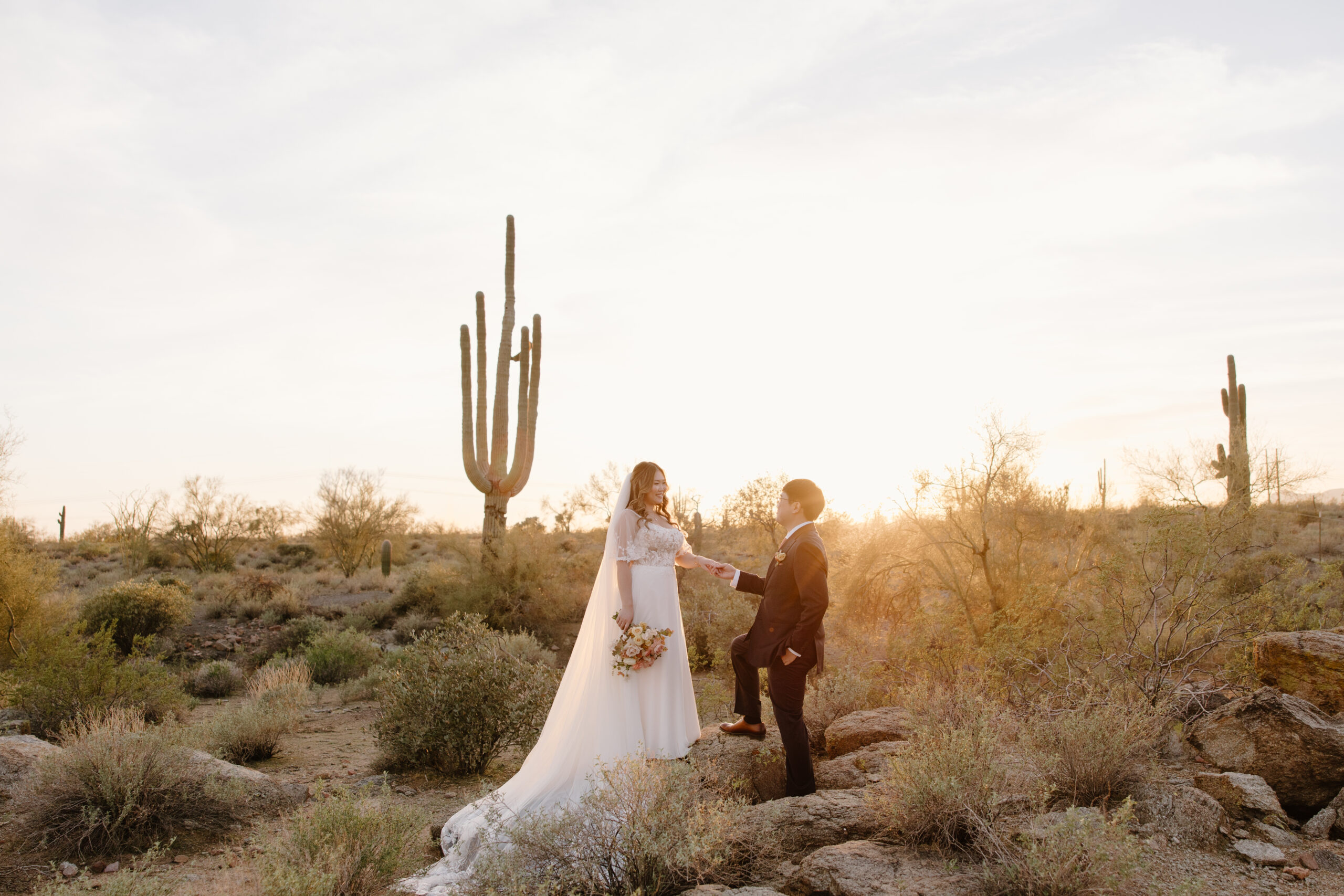 A bride in a white dress holds a bouquet, standing with a groom in a suit at sunset in a desert landscape with cacti, conveying romance and serenity.