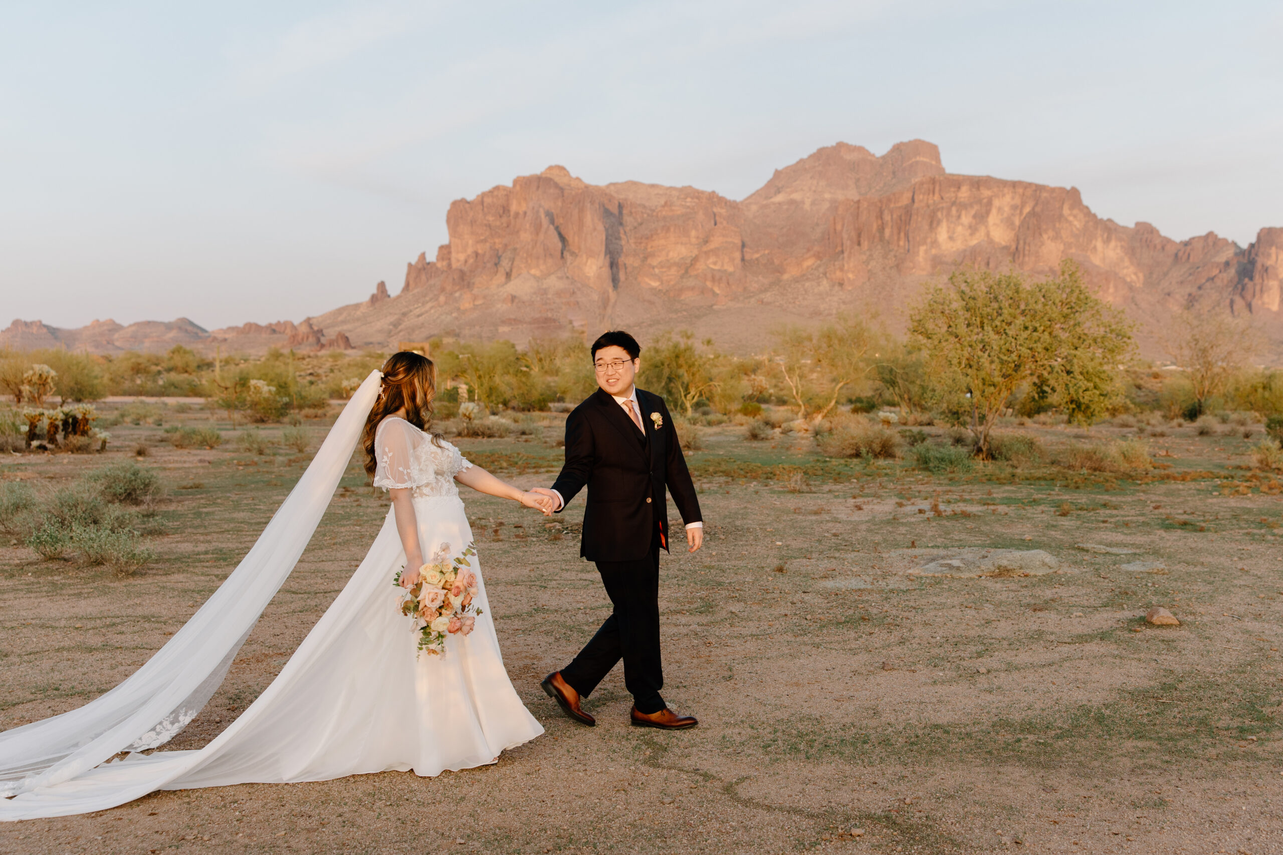 A couple walking hand in hand across the Arizona desert at The Paseo, Arizona, the bride’s veil flowing behind her like a ribbon in the warm desert breeze.