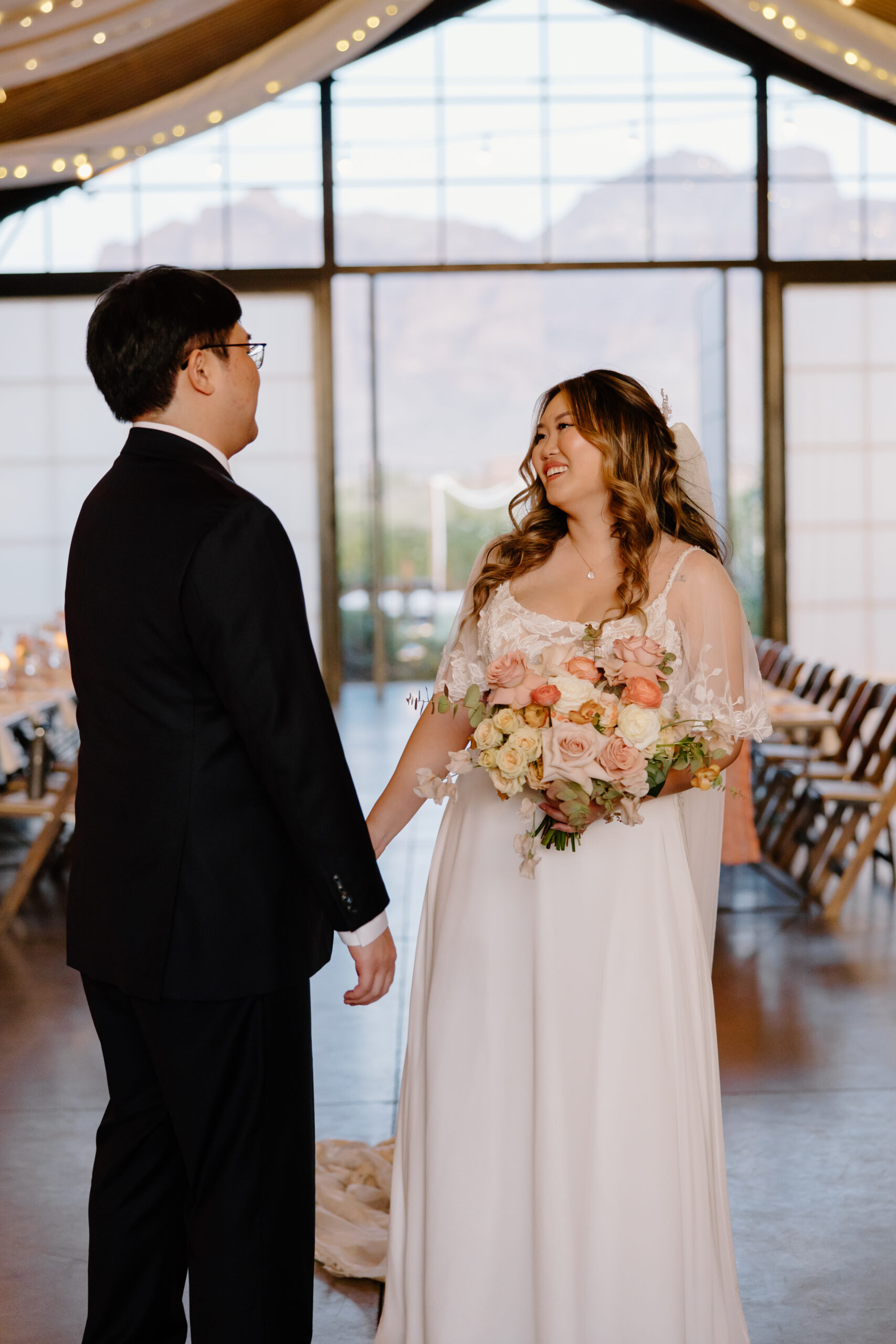 A bride in a white dress holds a bouquet and smiles at a groom in a dark suit inside a venue with large windows and mountain views.
