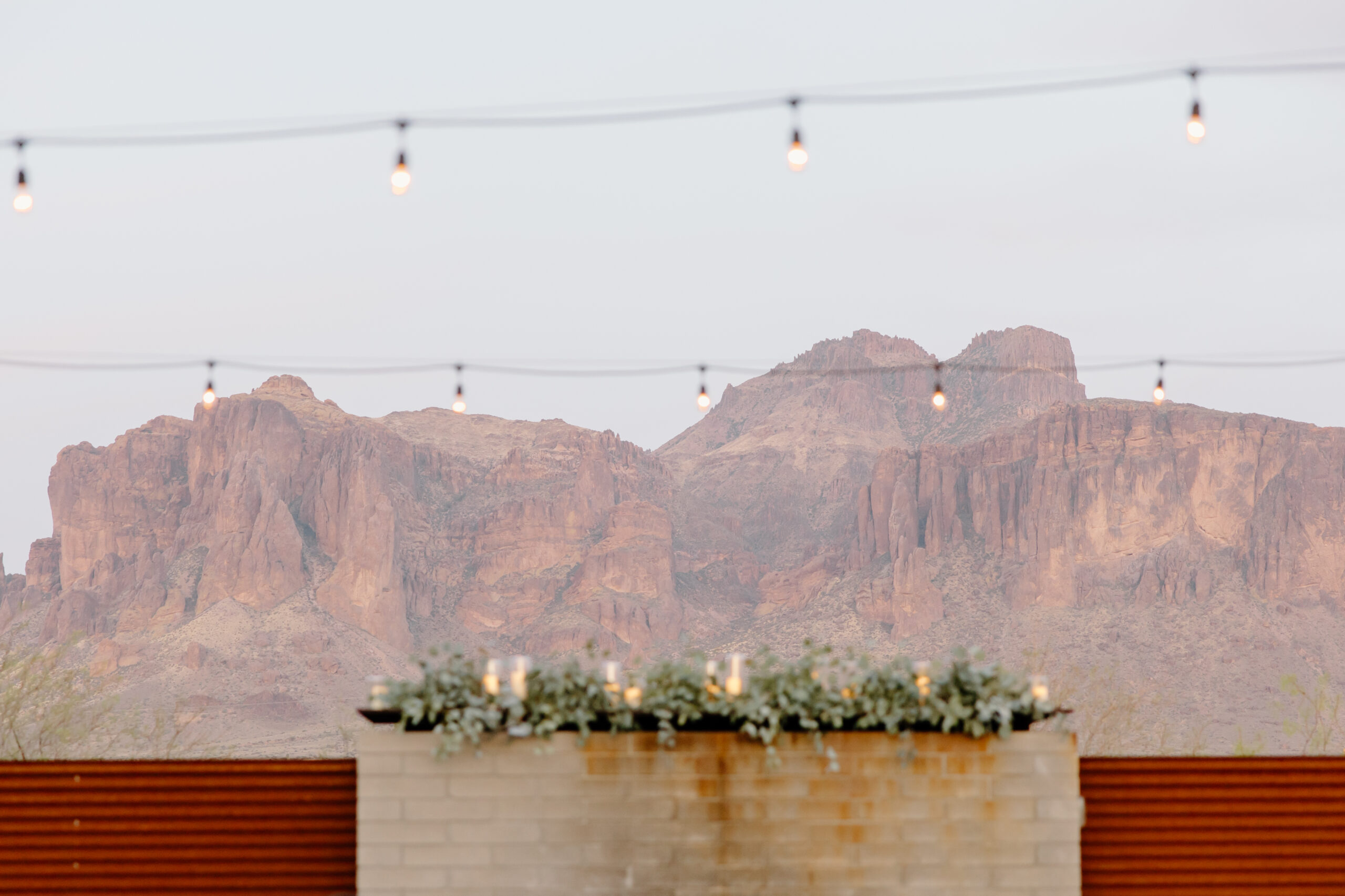 String lights hang above a brick wall adorned with greenery, set against a backdrop of towering, sunlit desert mountains, creating a serene atmosphere at The Paseo, Arizona.