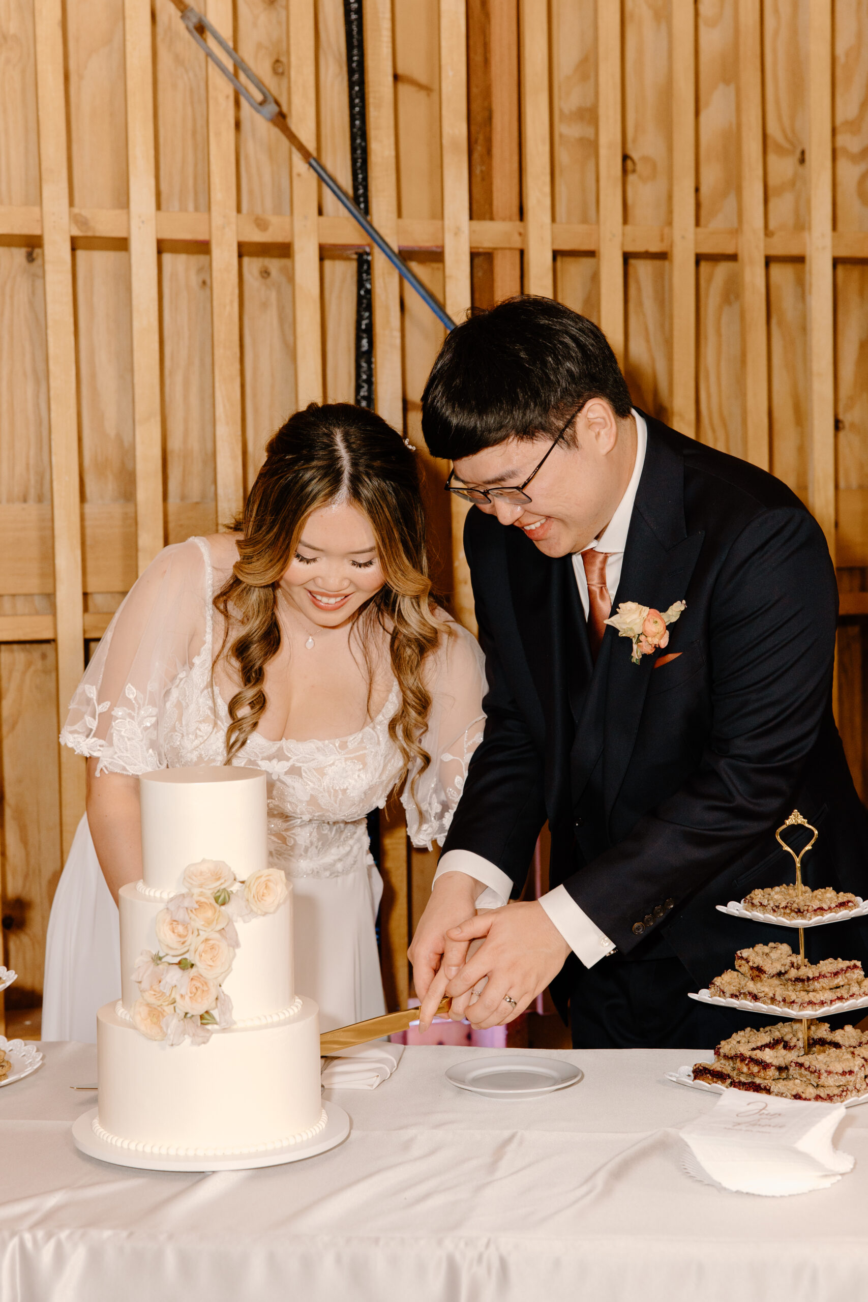 Bride and groom joyfully cut a tiered white wedding cake adorned with pastel flowers, standing in a wooden venue.