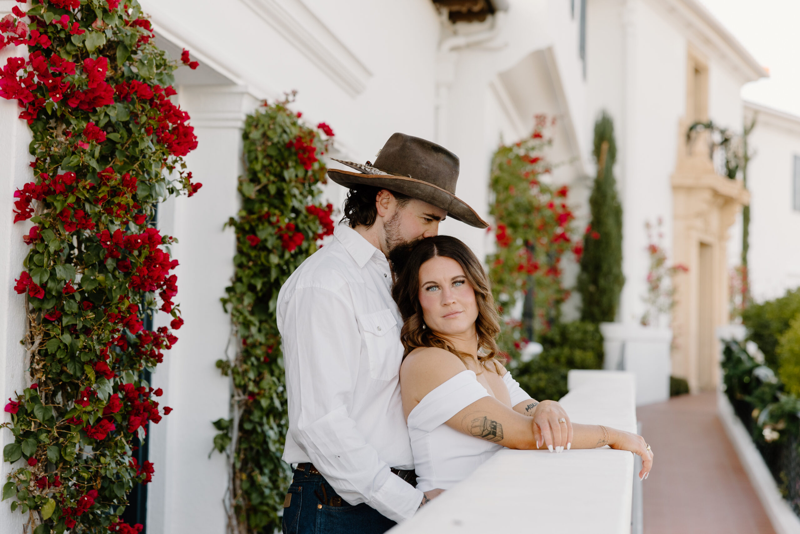 Couple walking hand in hand outside a white Spanish-style building with red flowers and black iron railings, a charming backdrop often featured among Arizona proposal locations.
