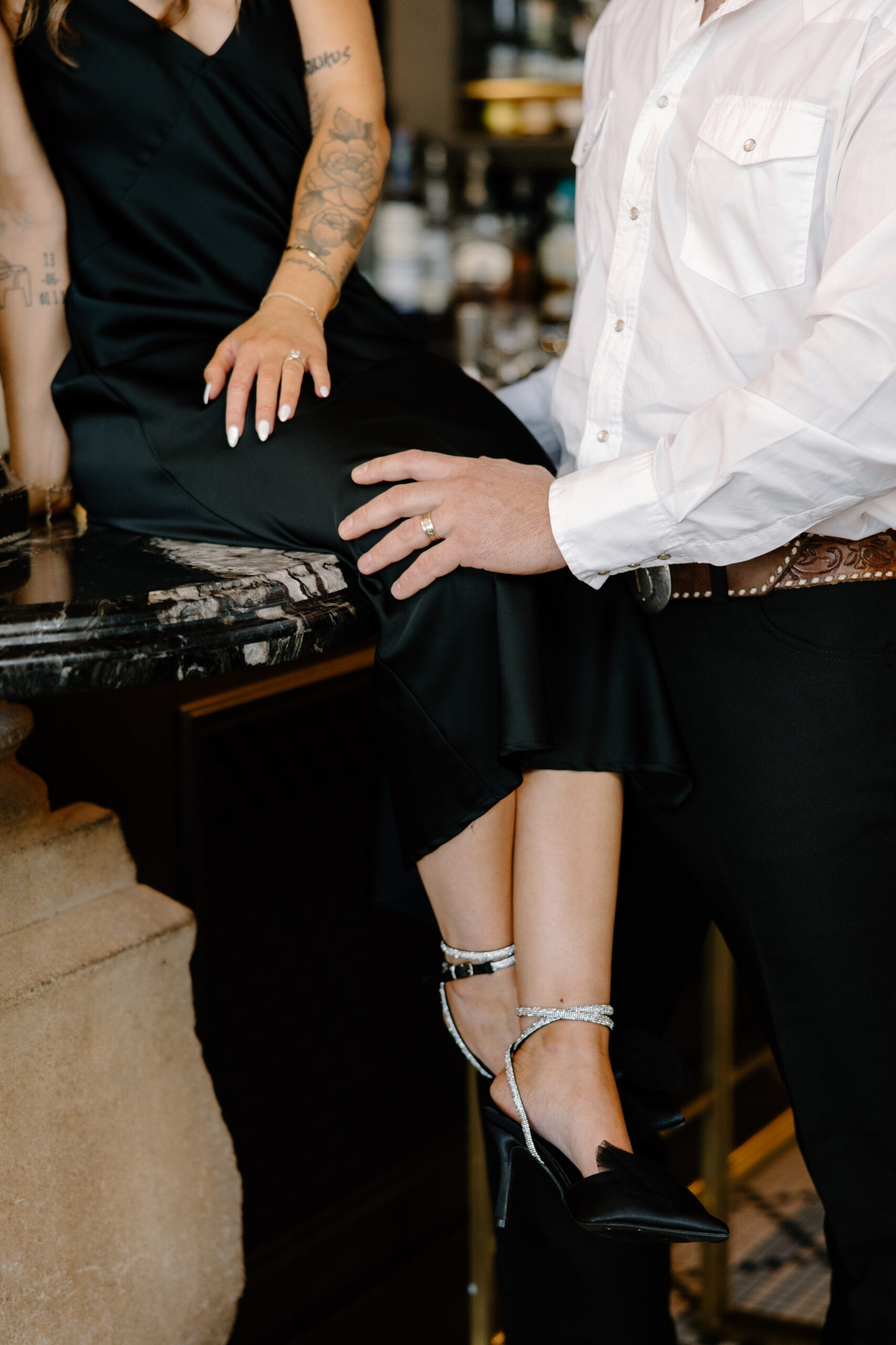 Detail shot of a couple dressed in black and white leaning against a bar, highlighting their hands, wedding bands, and elegant evening style.
