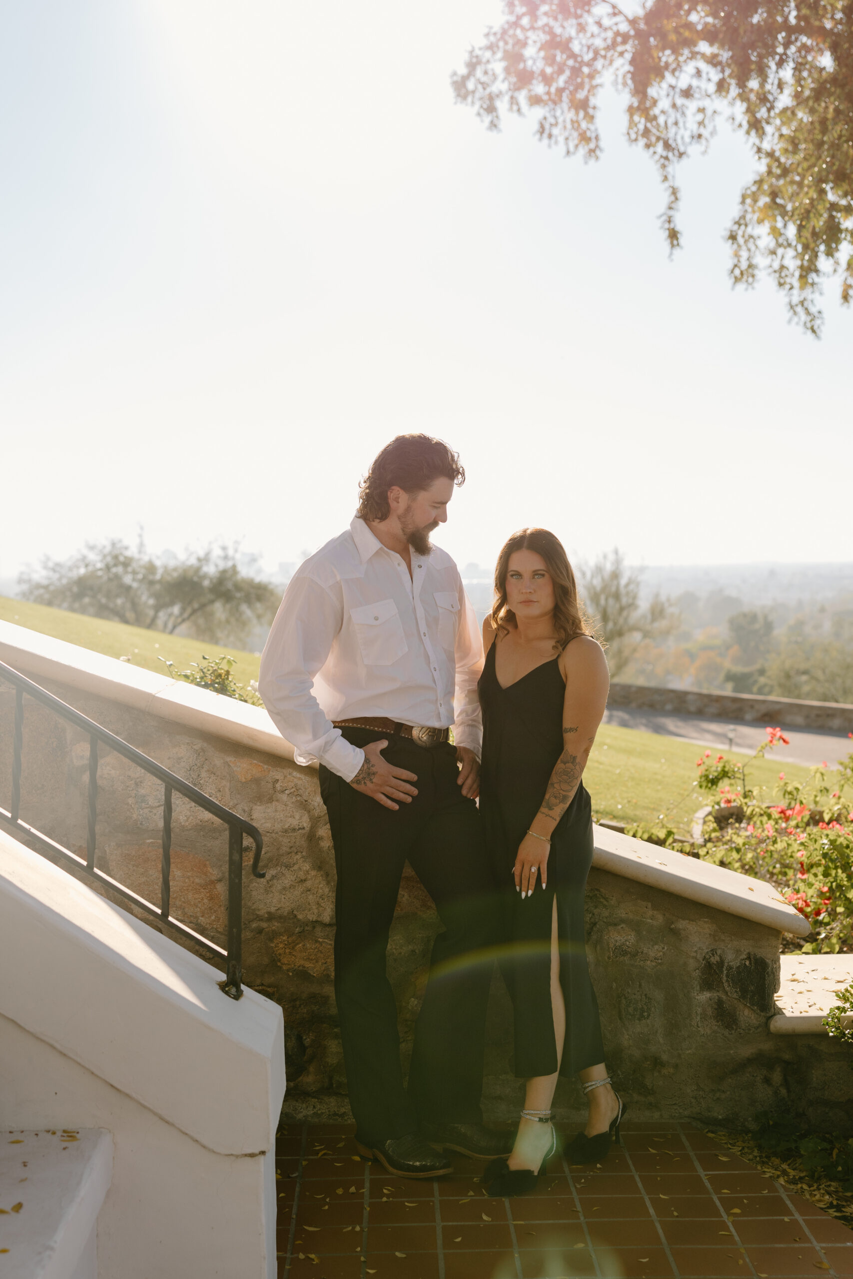 Couple standing together on a sunlit terrace, dressed in black and white with soft golden light wrapping around them.