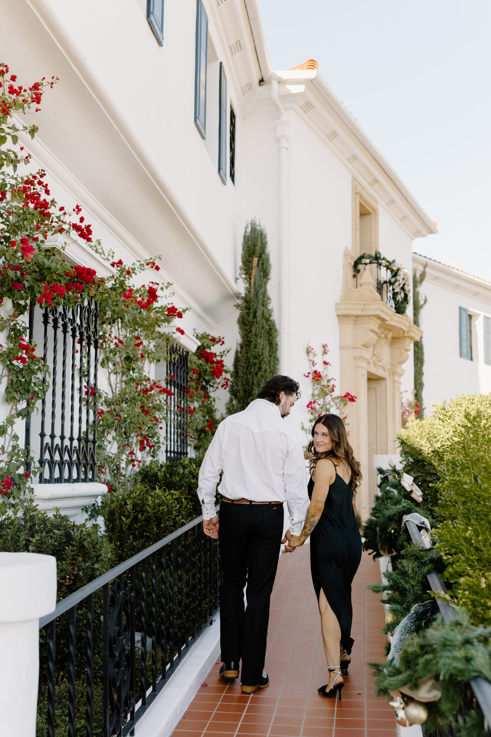 Couple walking hand in hand outside a white Spanish-style building with red flowers and black iron railings, a charming backdrop often featured among Arizona proposal locations.