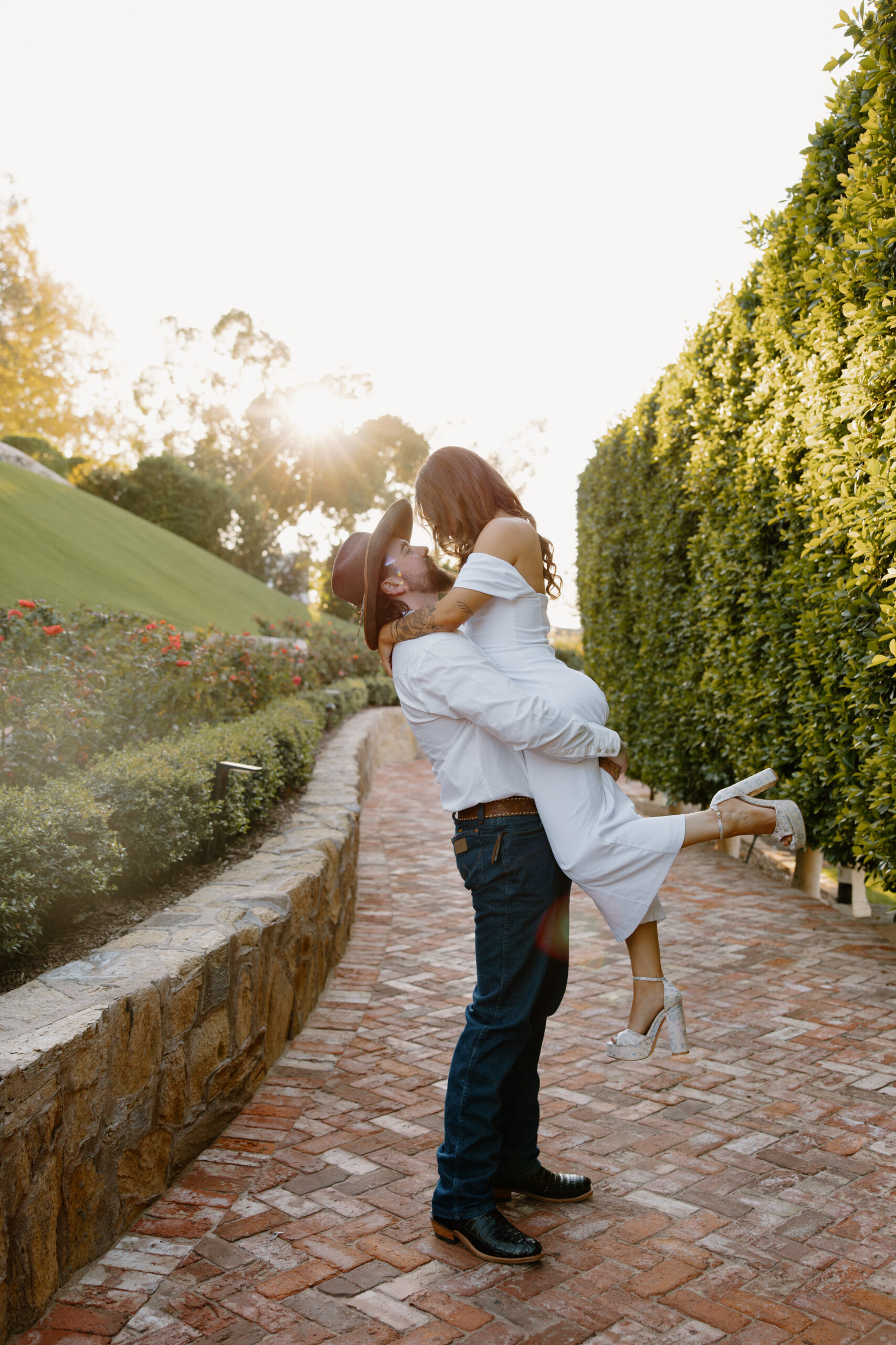 Couple embracing on a brick pathway at golden hour, surrounded by manicured hedges and warm sunlight, creating a romantic moment at one of the most charming Arizona proposal locations.