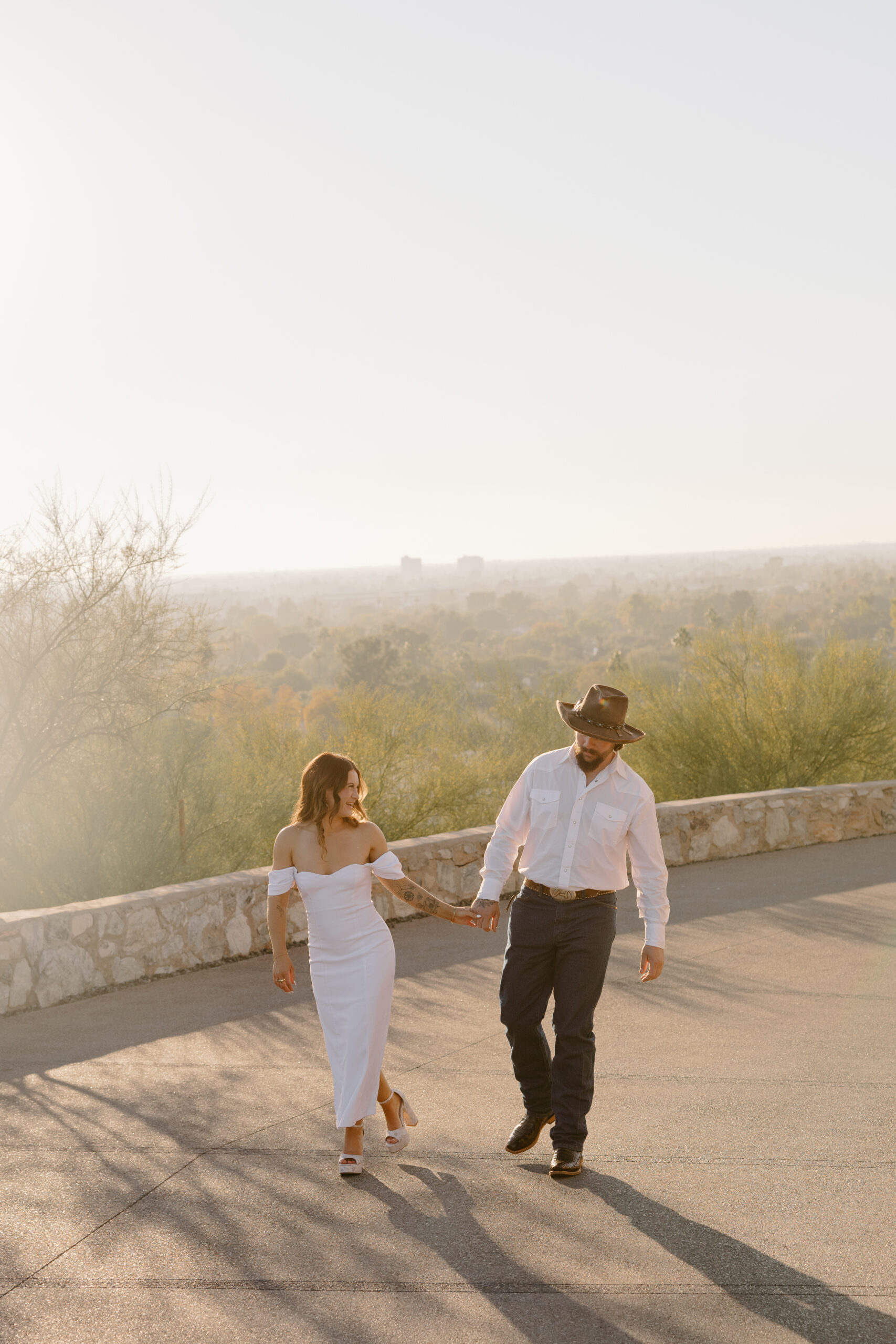 Couple strolling at golden hour on a scenic overlook with desert views and long shadows stretching across the pavement.