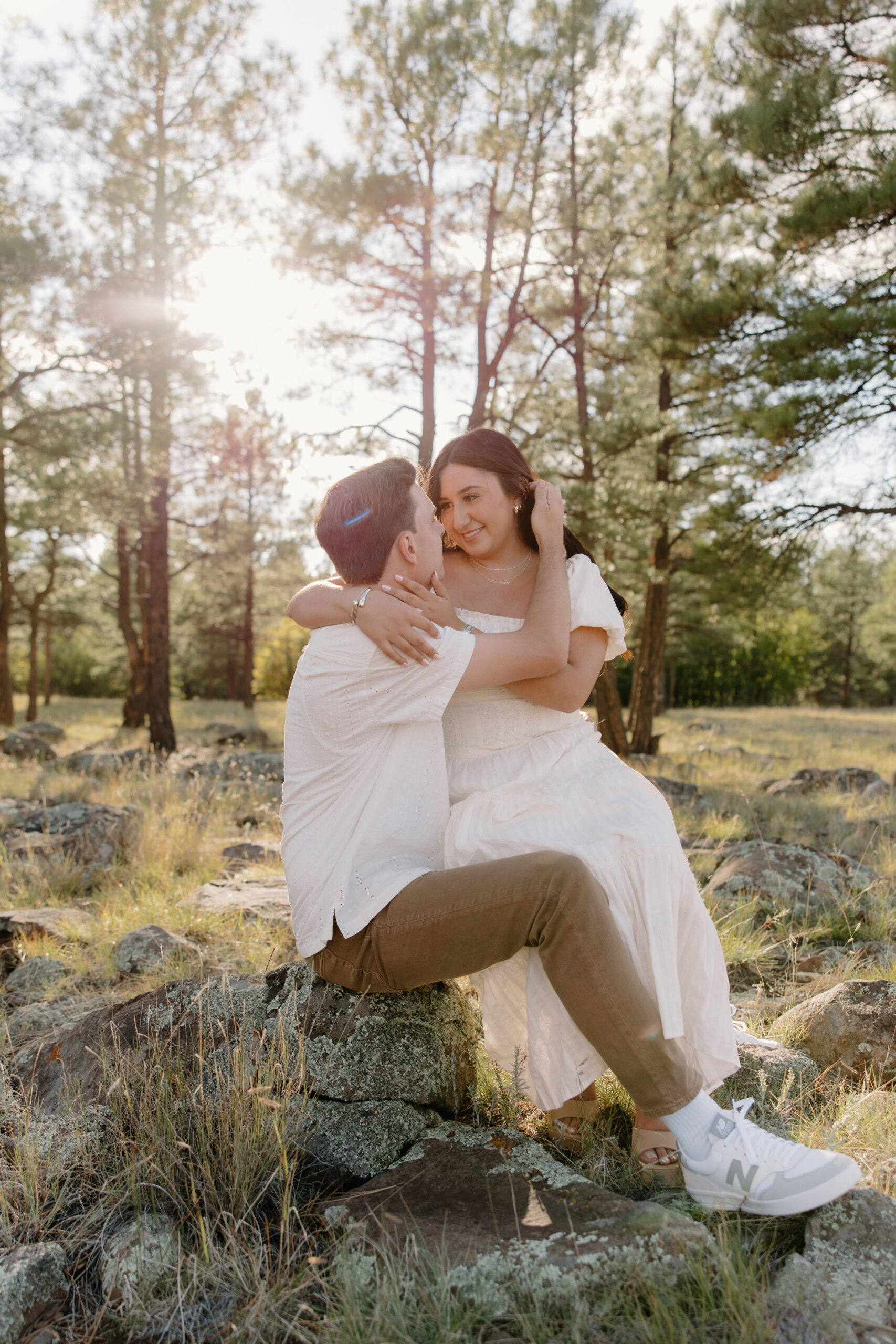 Bride sitting on her partner’s lap on sunlit rocks in a pine forest, smiling during an intimate engagement session in one of Arizona’s scenic proposal locations.