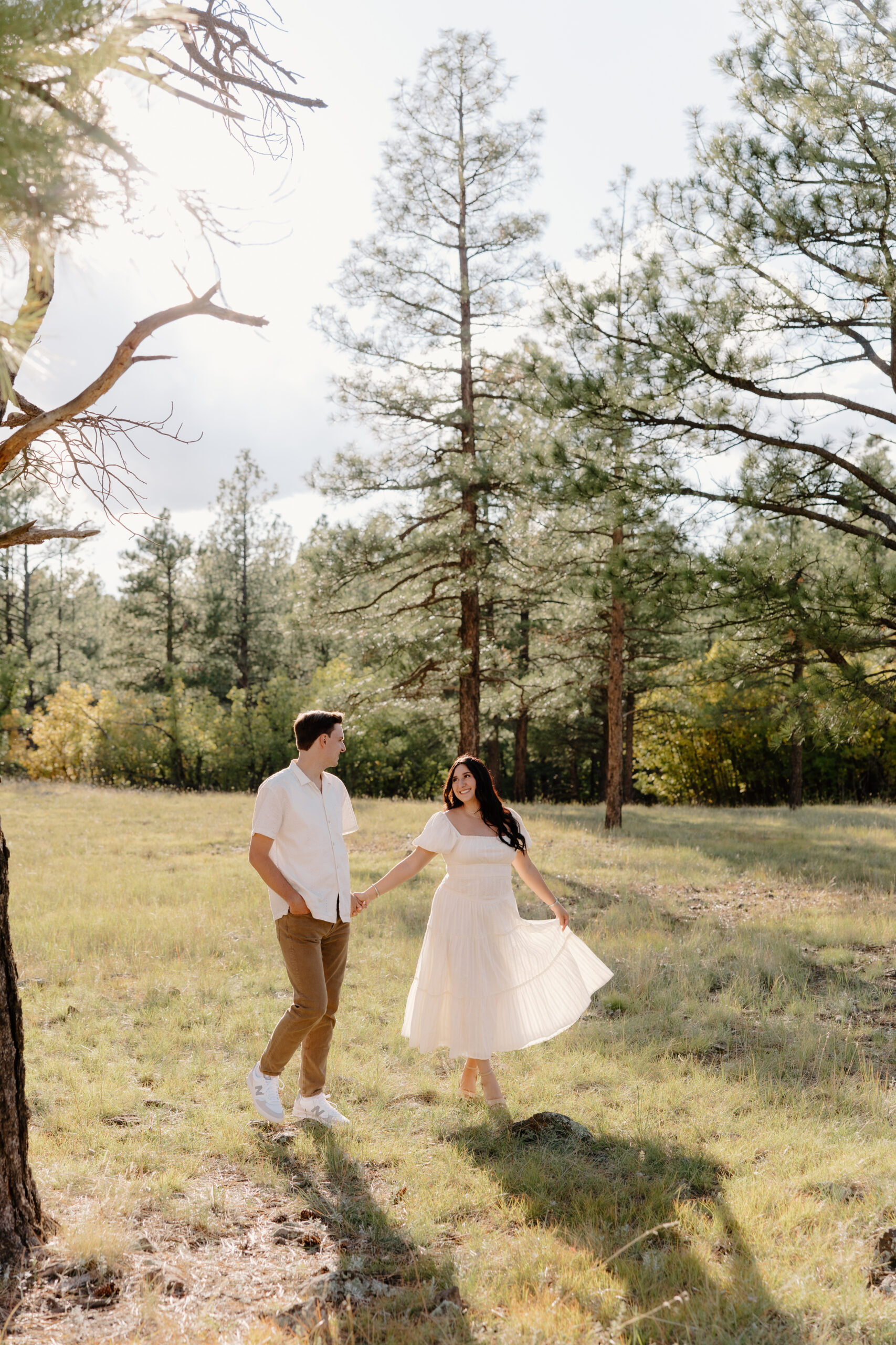 Joyful couple holding hands and twirling in a sunlit forest meadow with tall pine trees and soft golden light filtering through the branches.