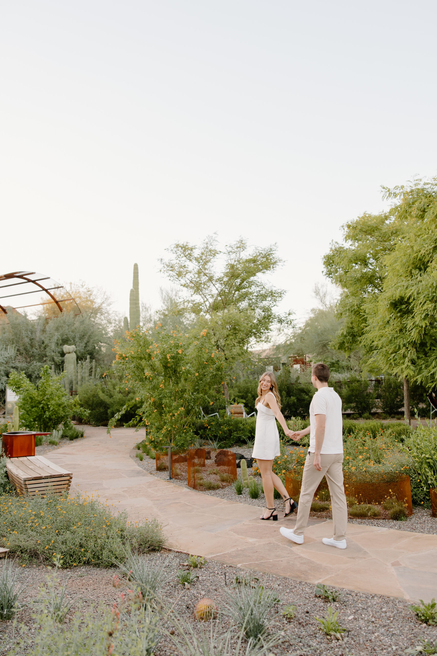 Couple walking hand in hand through a desert garden pathway filled with cacti and greenery, capturing a candid engagement moment at one of Arizona’s hidden proposal locations.
