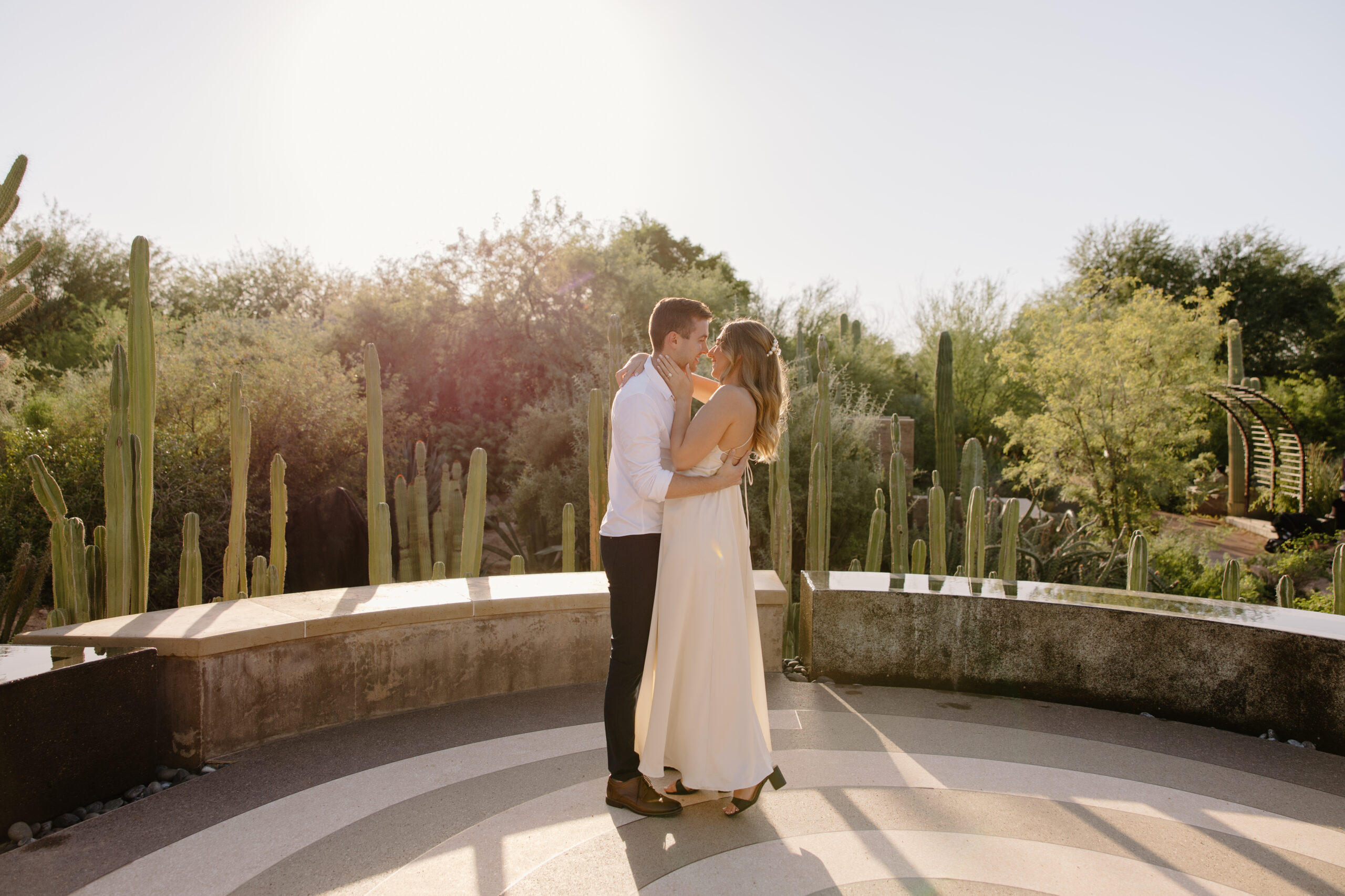 Couple sharing a kiss on a circular desert terrace surrounded by tall cacti and warm golden light, highlighting the beauty of elevated Arizona proposal locations.