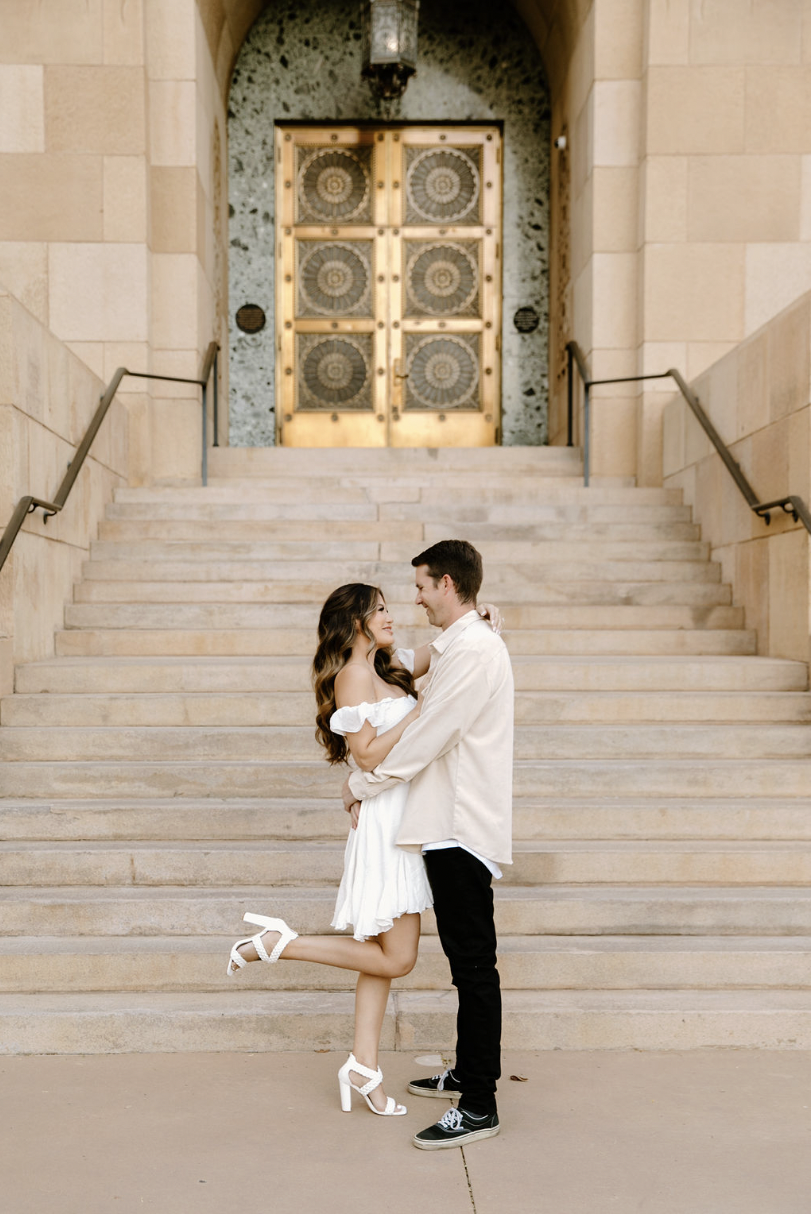 Couple embracing on the Phoenix City Hall steps, her white dress and heels popping against the warm stone architecture as they smile at each other.