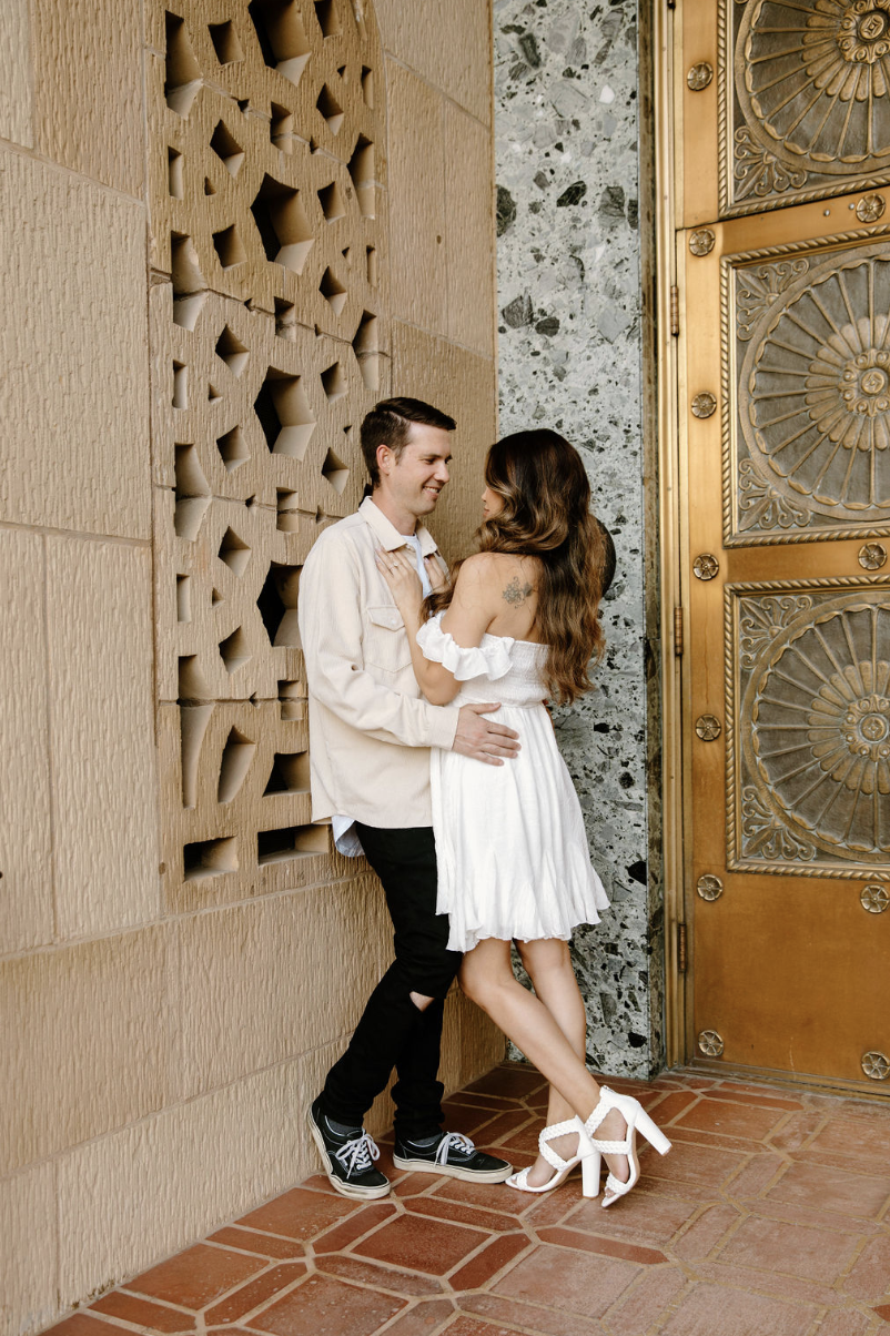 Couple leaning against a textured stone wall beside decorative bronze doors, sharing a quiet, romantic moment in downtown Phoenix.