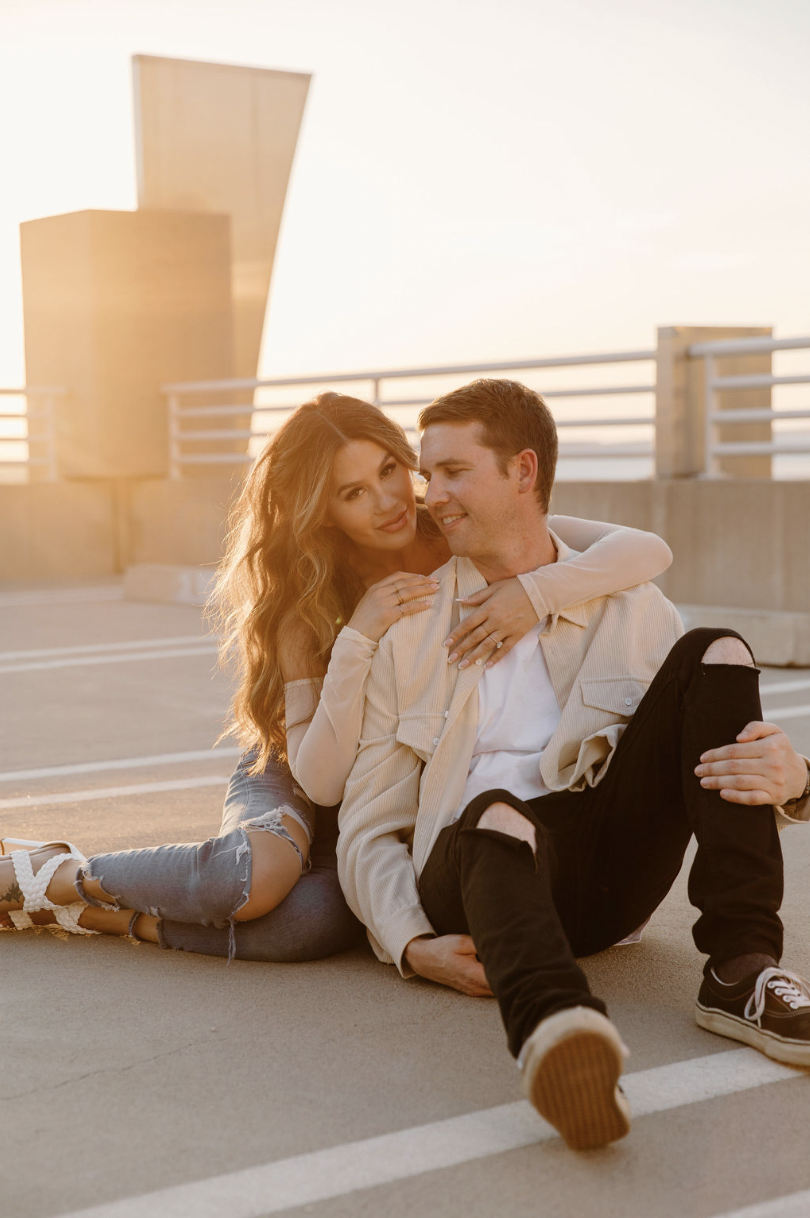 Engaged couple sitting on a downtown rooftop parking garage at sunset, wrapped up together with soft golden light behind them.
