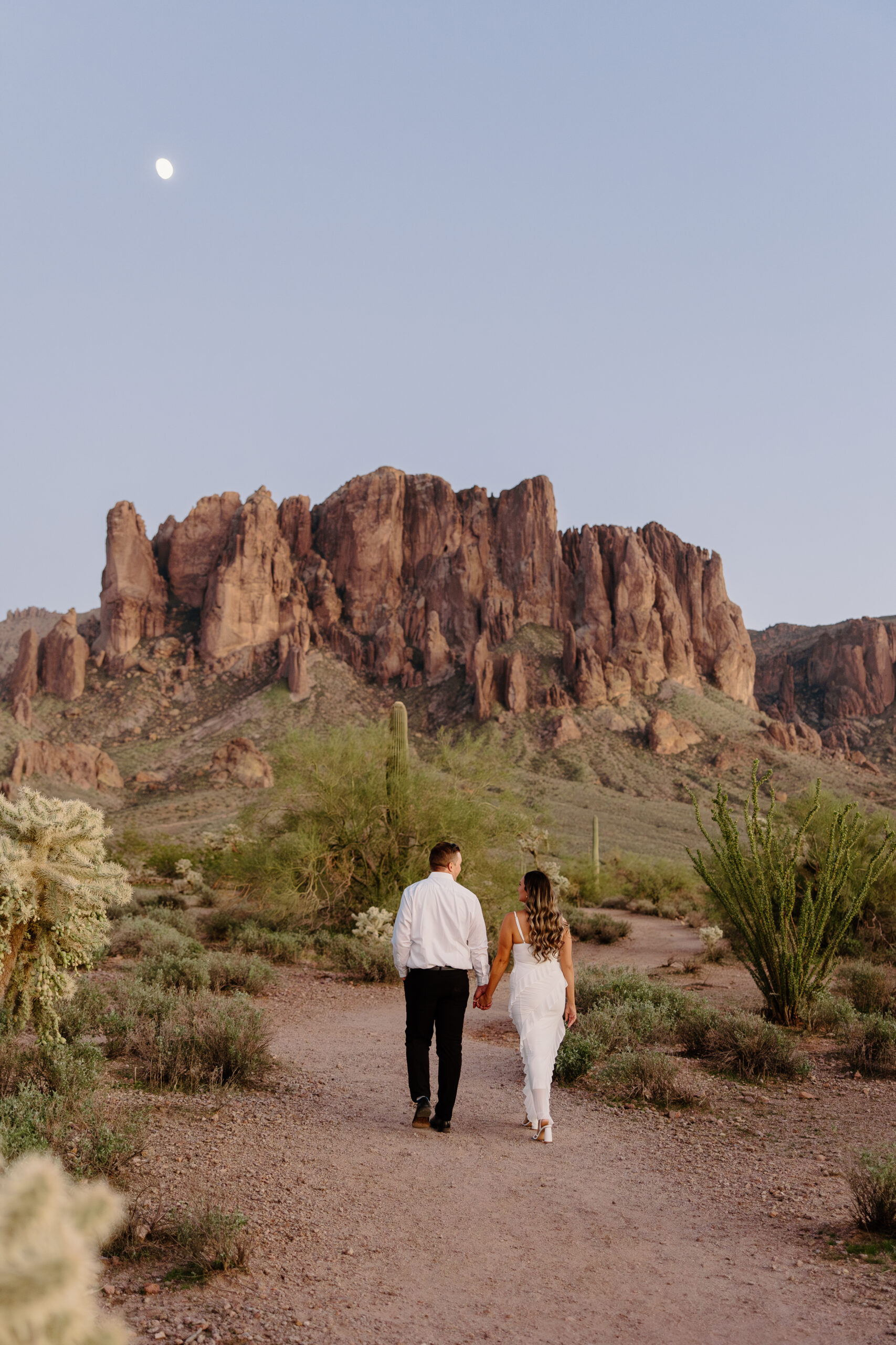 Couple walking hand in hand through the desert at sunset with dramatic red rock cliffs and saguaro cacti in the background at one of the most scenic Arizona proposal locations.