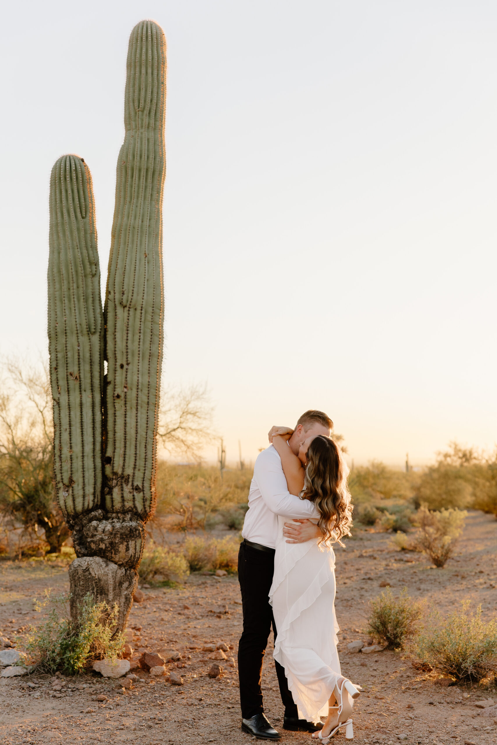Couple kissing at sunset beside a towering saguaro cactus in the desert, golden light wrapping around them during a romantic moment at one of the most iconic Arizona proposal locations.
