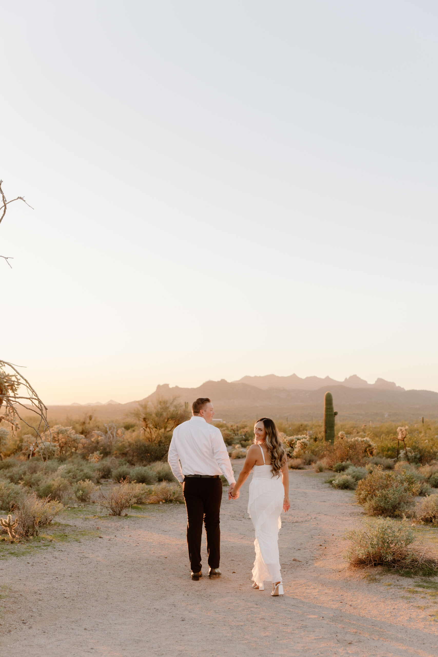 Newly engaged couple walking hand in hand down a desert path at sunset with mountains in the distance, showcasing the natural beauty of Arizona proposal locations.
