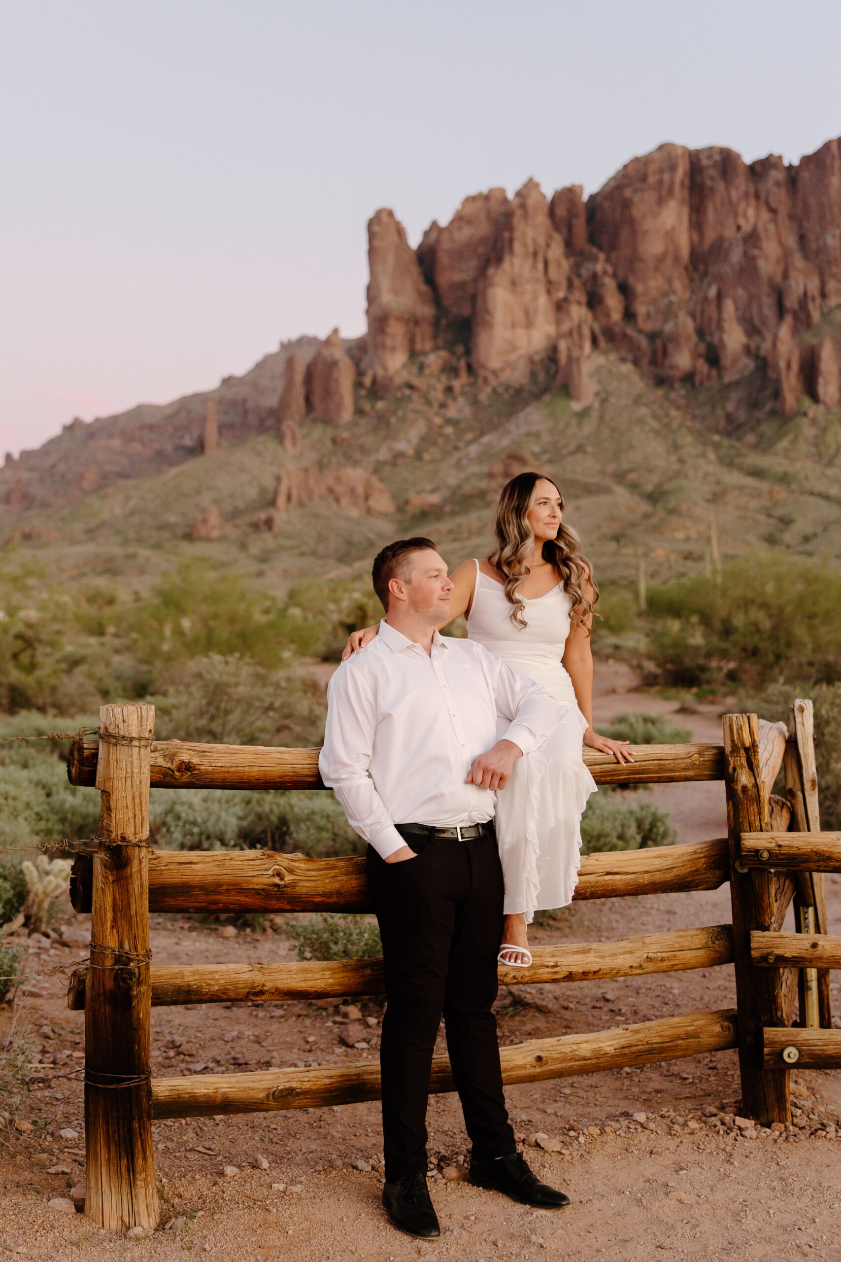 Couple posing on a rustic wooden fence with the Superstition Mountains glowing behind them at sunset, a stunning backdrop for Arizona proposal locations.