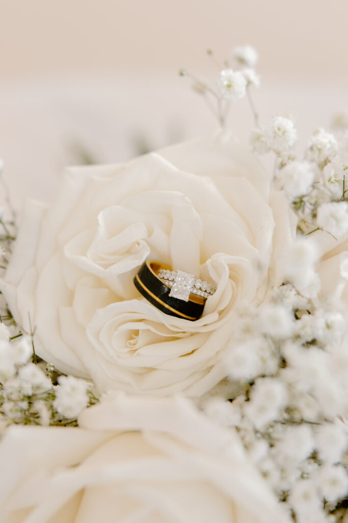 Close-up of the bride’s engagement ring and wedding band resting inside a white rose surrounded by delicate baby’s breath.