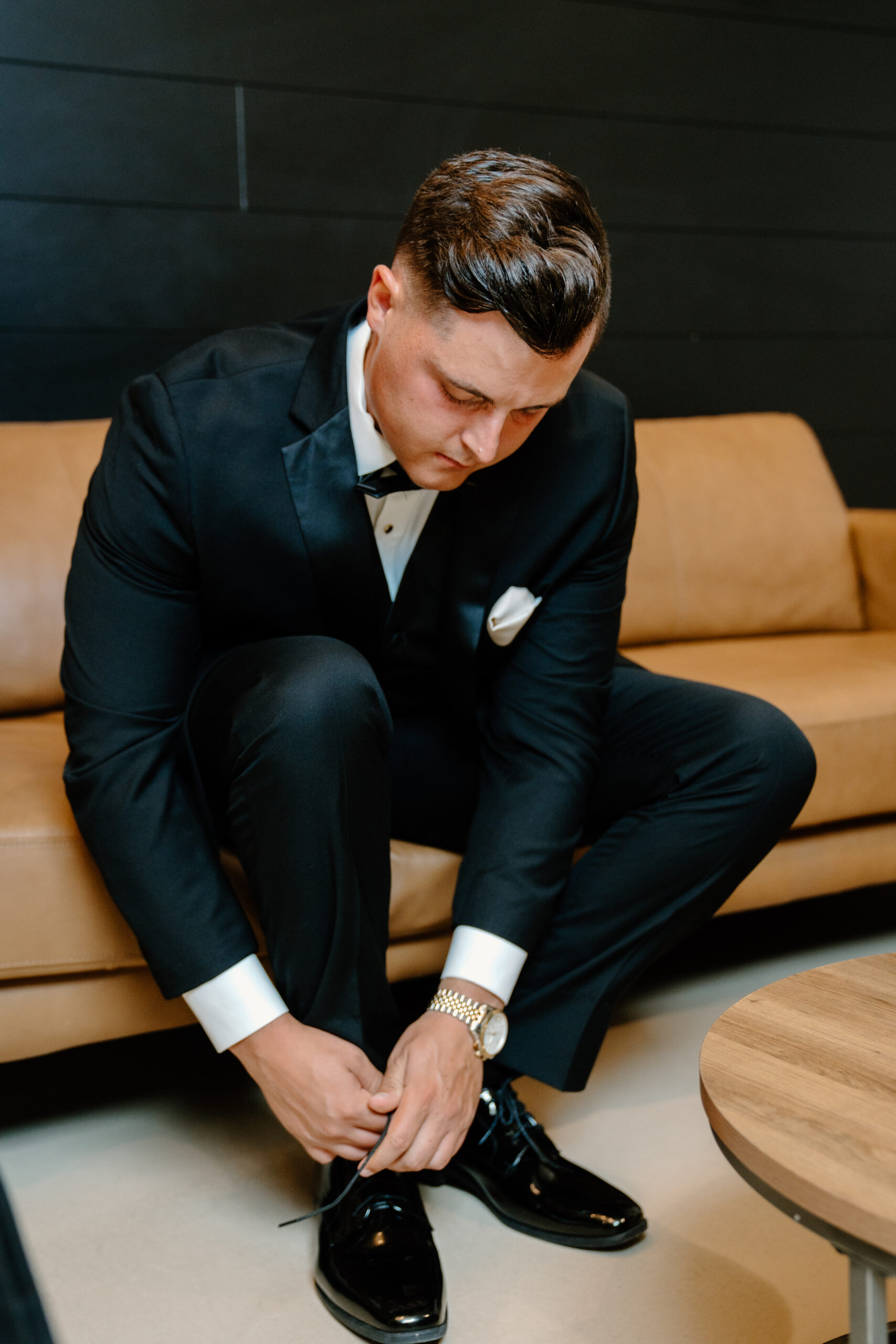 Groom sitting on a leather sofa tying his dress shoes while getting ready for the wedding day.