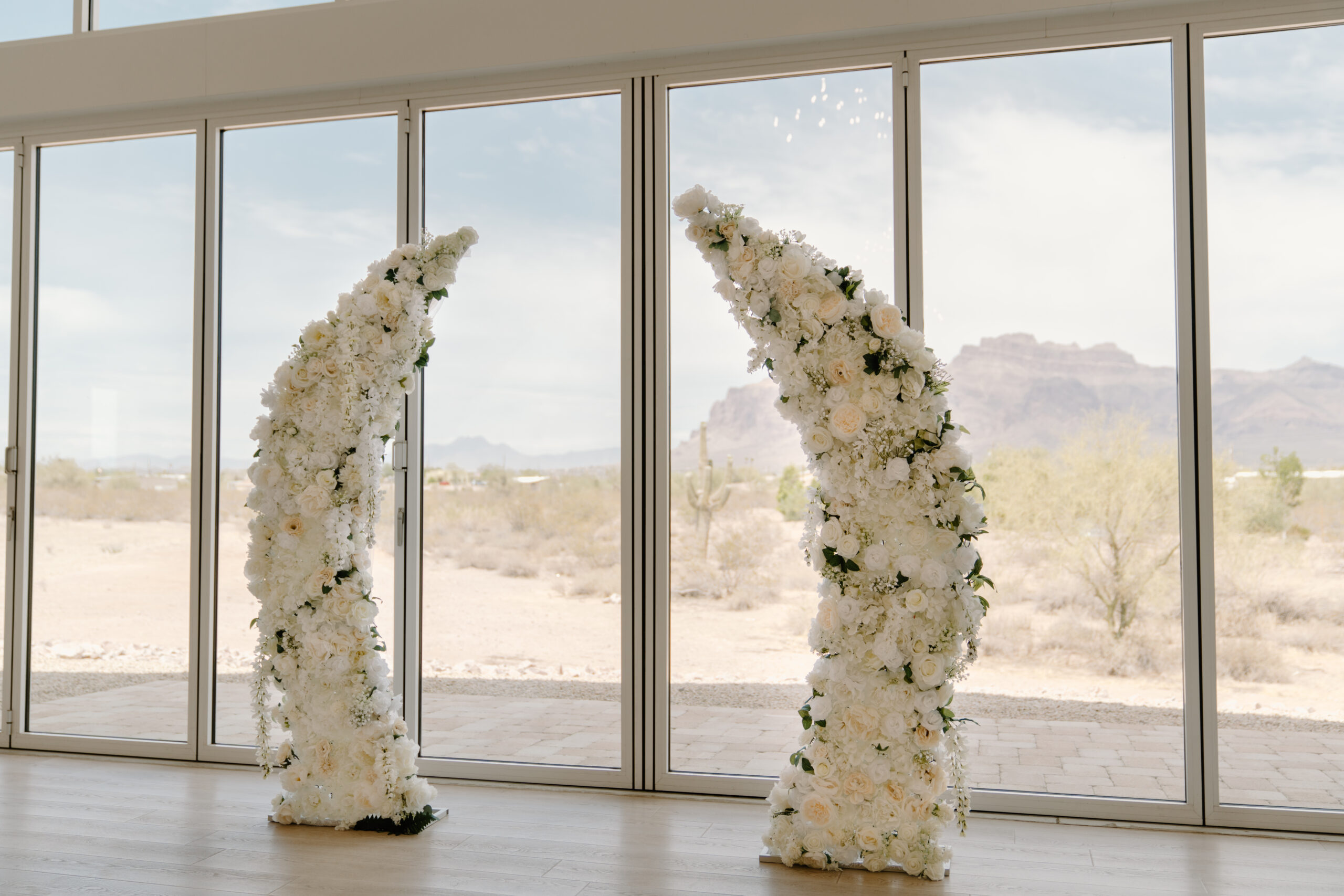 Modern white floral ceremony arches positioned in front of floor-to-ceiling windows overlooking the desert at a Desert View wedding.