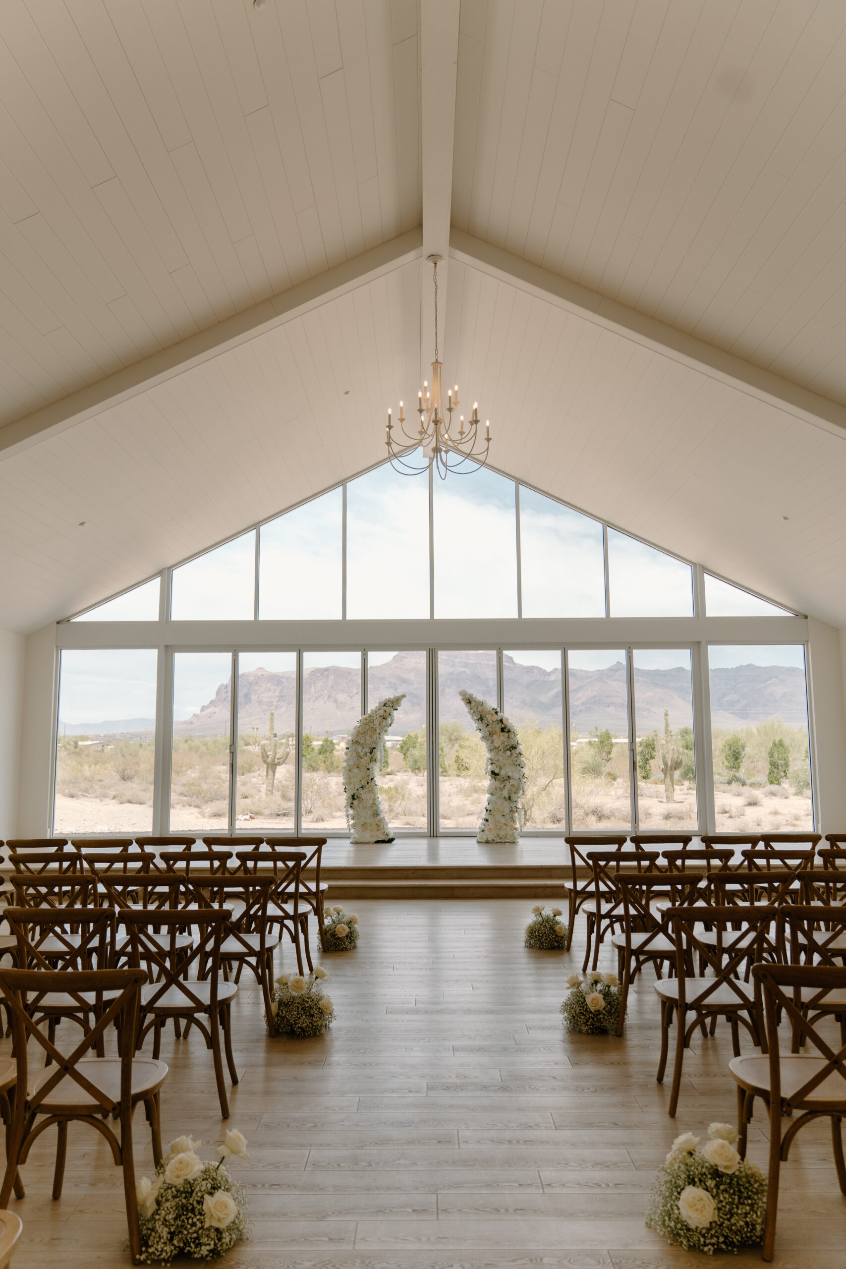 Bright ceremony space with wooden chairs, a floral arch, and panoramic windows overlooking desert mountains at a Desert View wedding venue.