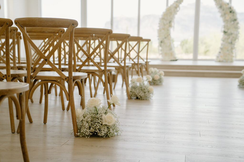 Wooden ceremony chairs with white floral aisle arrangements inside the bright chapel ceremony space.