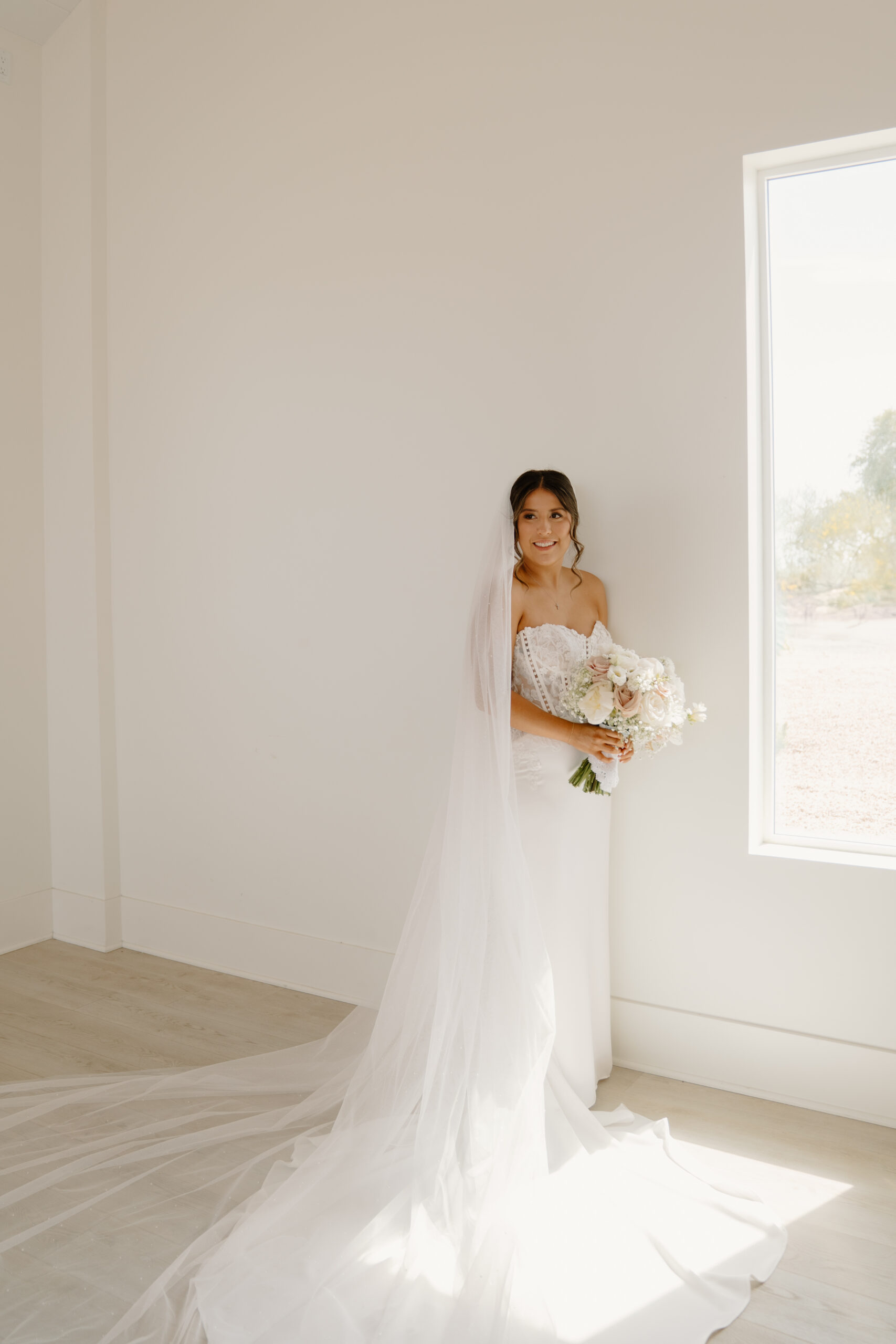 Bride standing near a bright window holding her bouquet with her long veil flowing across the floor inside the chapel.