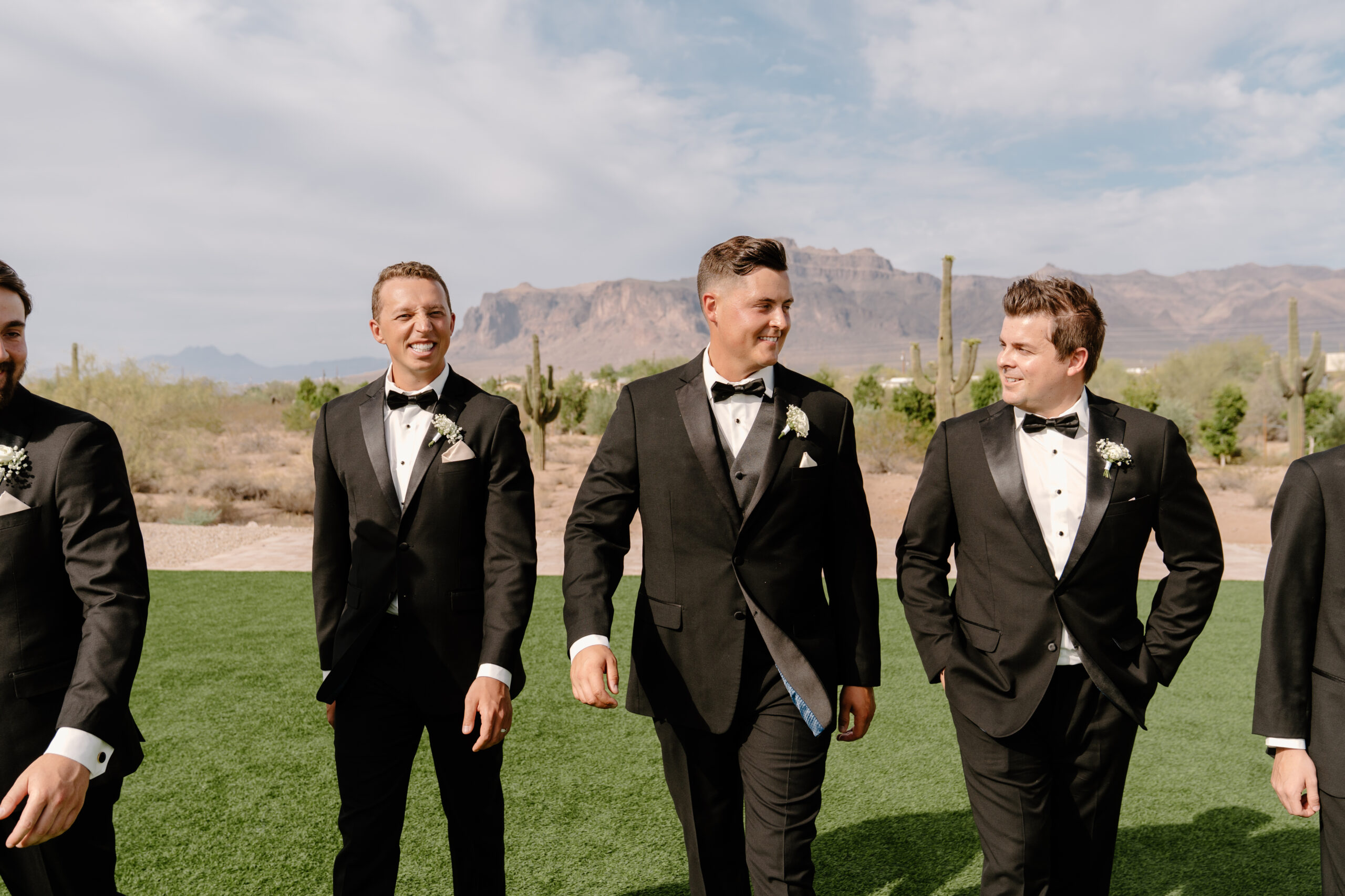 Groom and groomsmen walking together outdoors in black tuxedos with desert mountains and saguaros in the background.