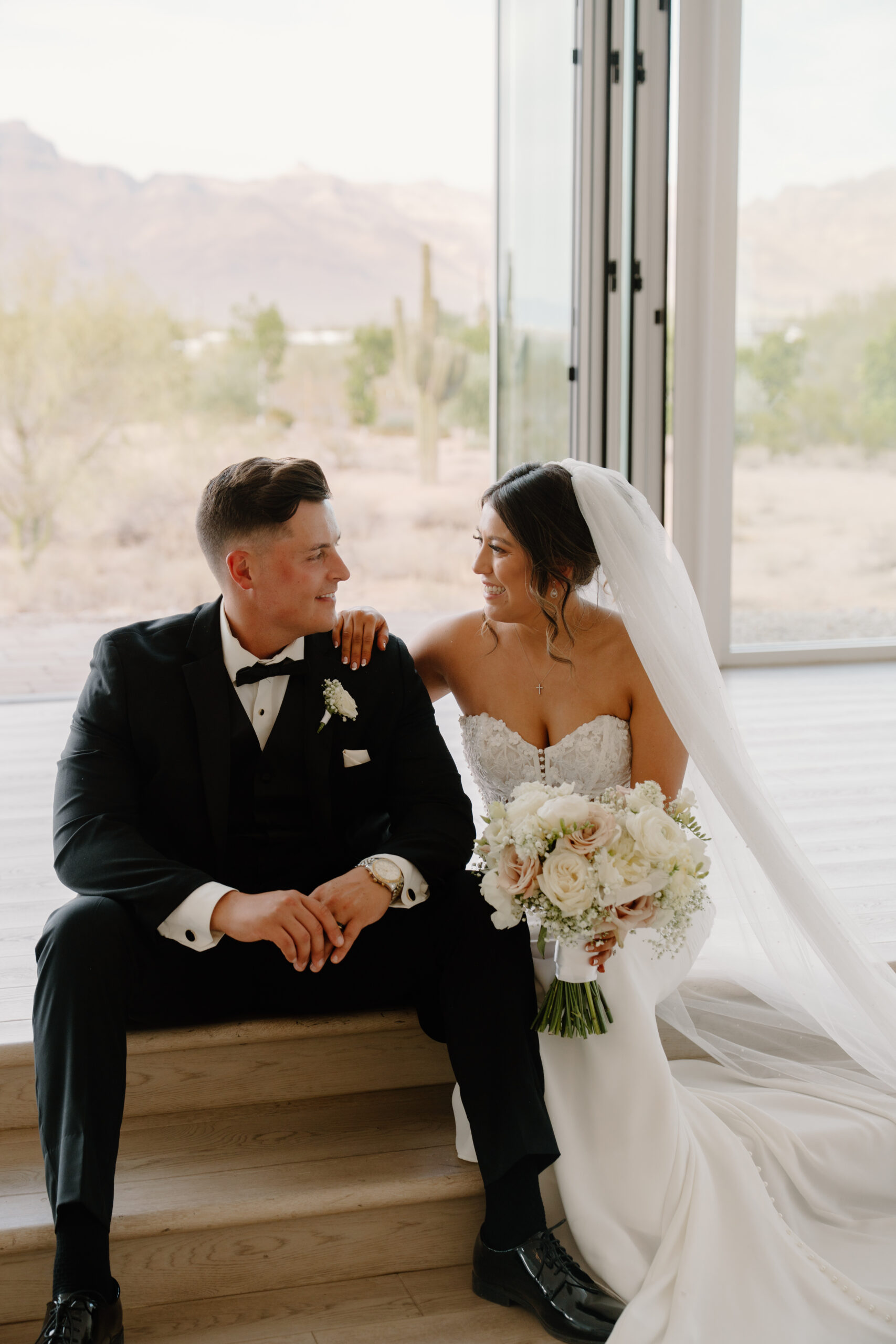 Bride and groom sitting together on wooden steps inside the bright chapel with desert views through large windows.