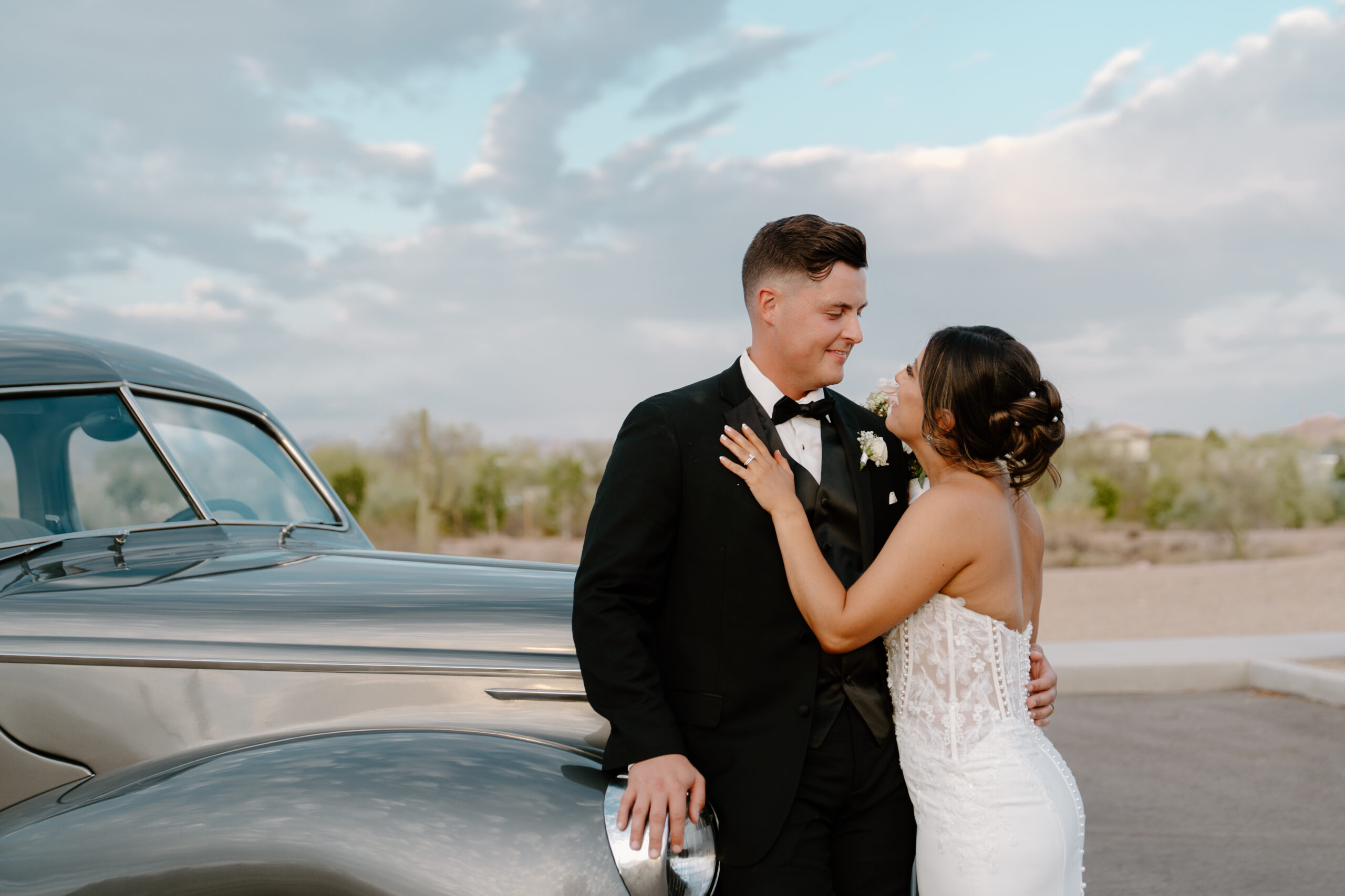 Bride and groom embracing beside a vintage car with desert mountains and soft evening light.