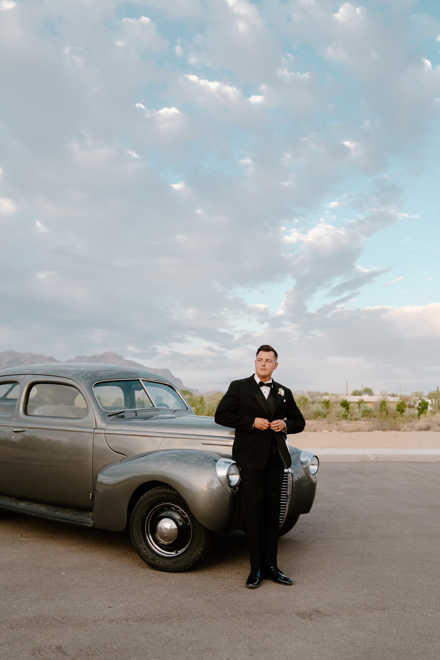 Groom standing beside a vintage silver car with desert mountains and dramatic sky in the background during a Desert View wedding.
