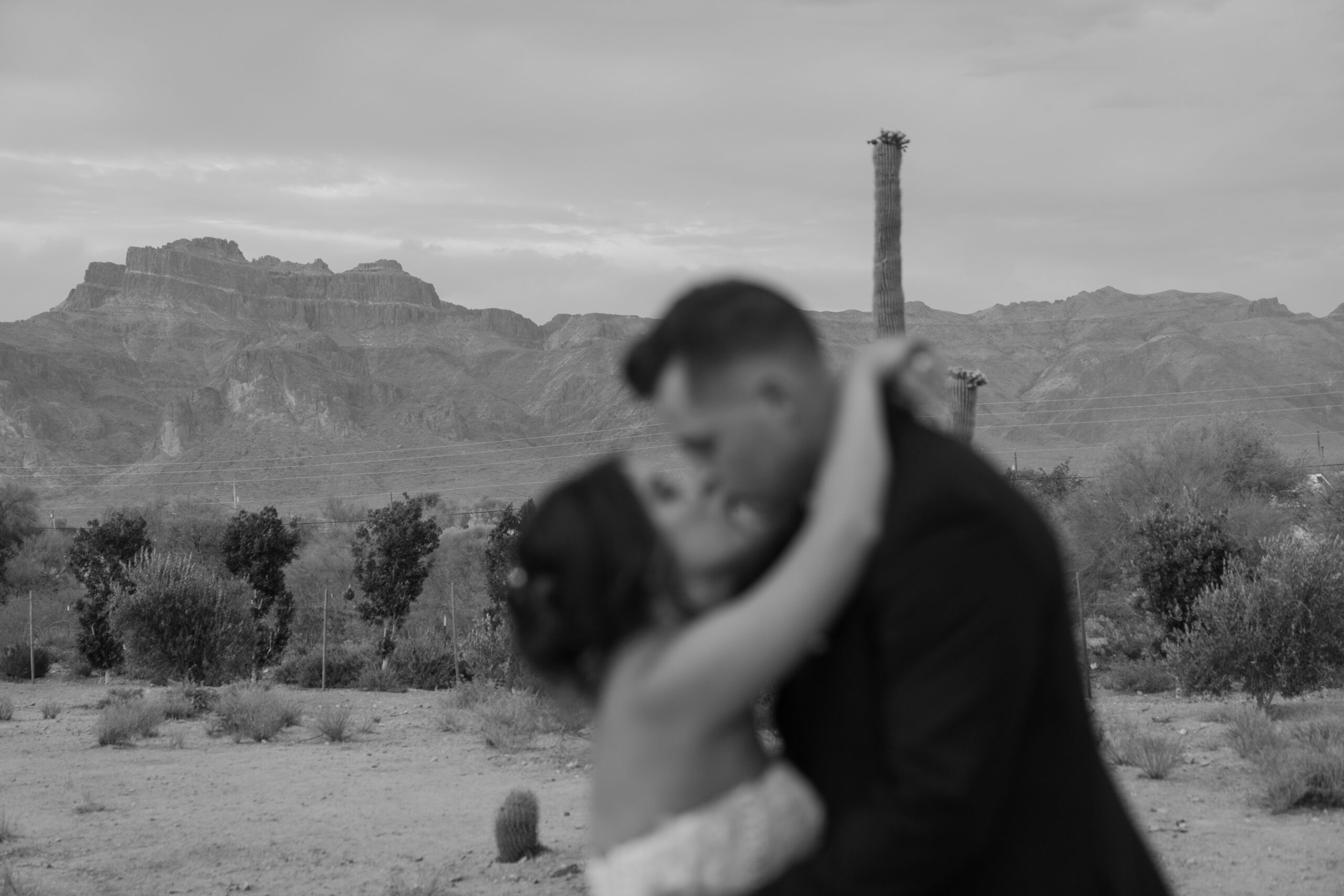 Black and white portrait of the bride and groom embracing with the Superstition Mountains and desert landscape behind them.