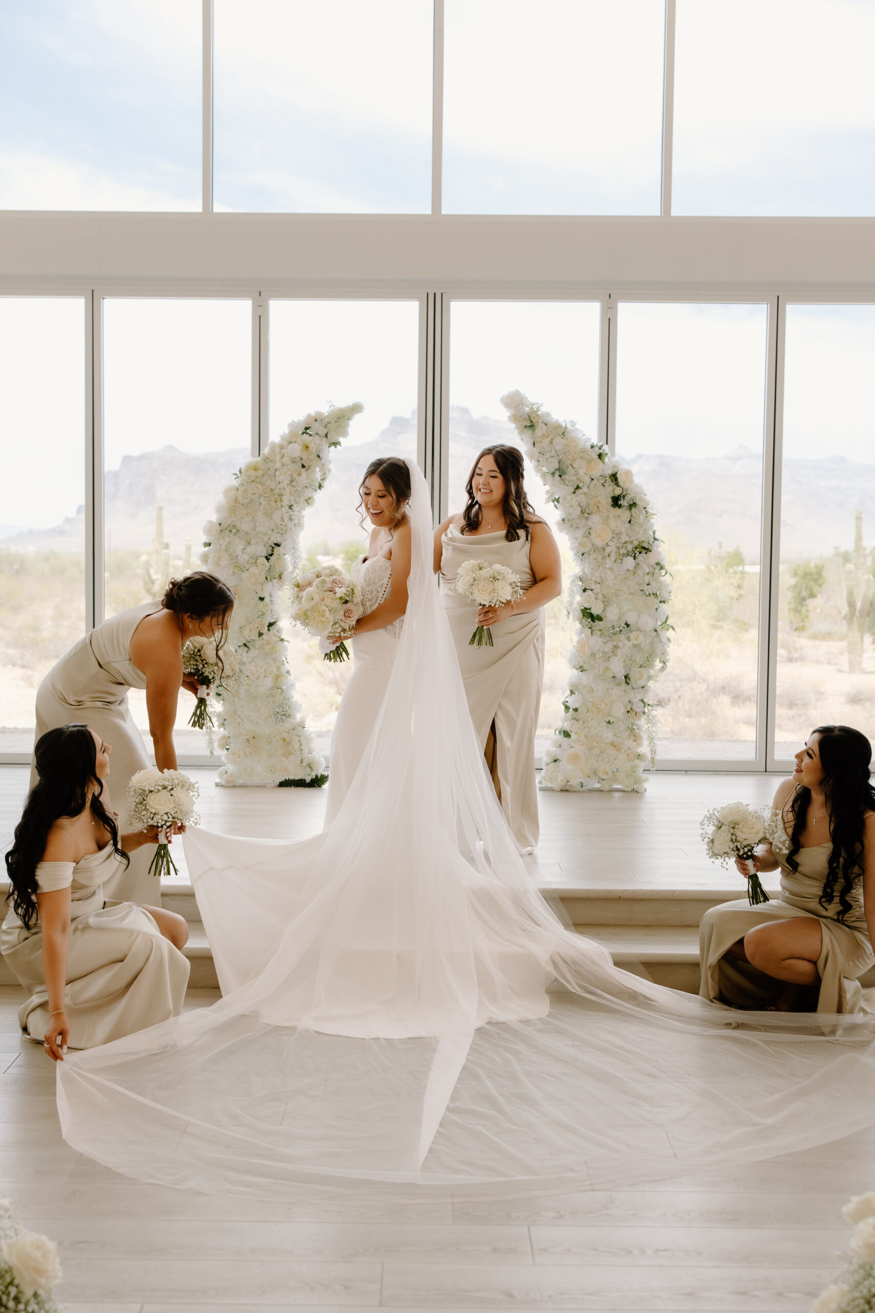 Bride standing with bridesmaids while they arrange her long veil and train in front of floral ceremony arches at a Desert View wedding.