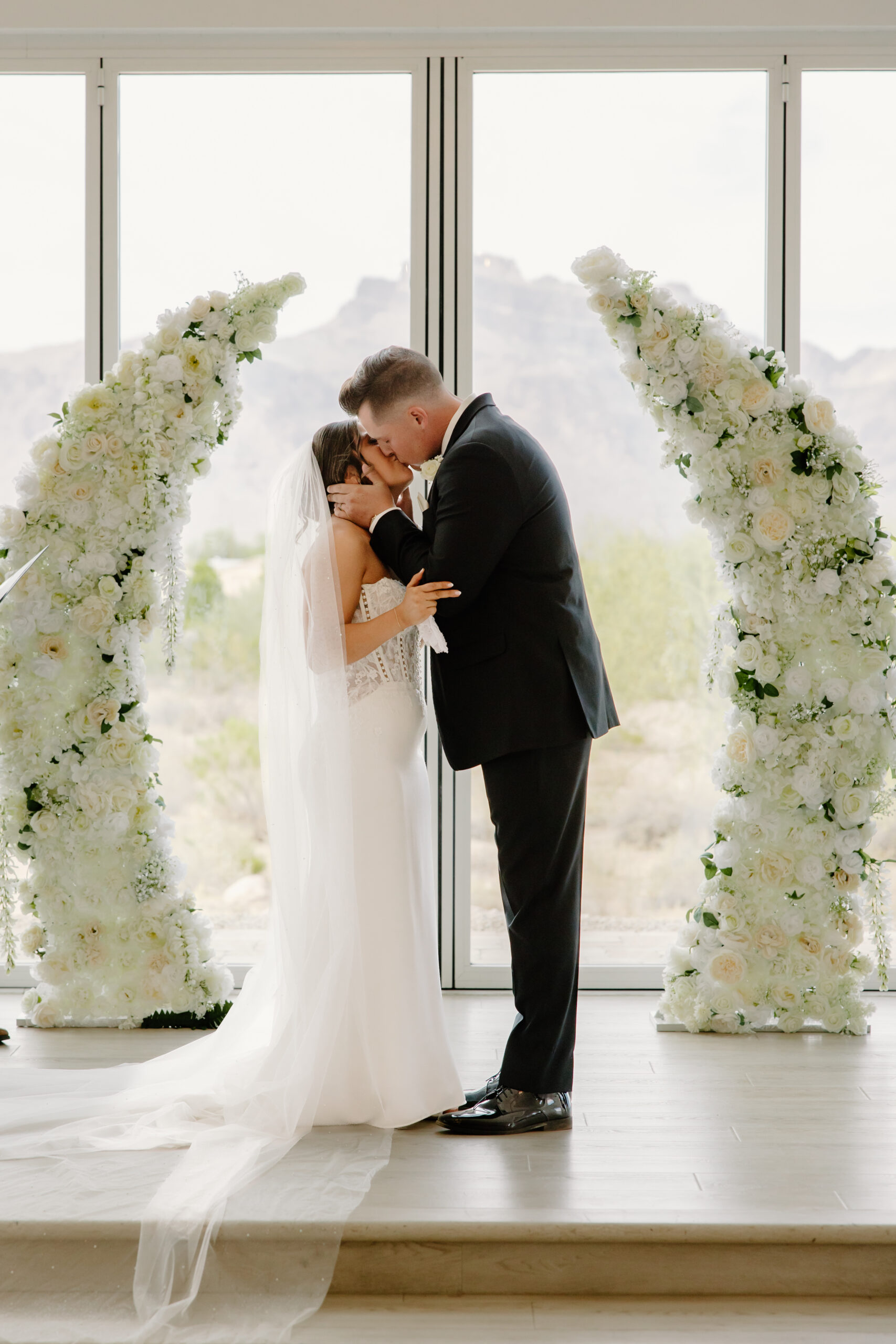 Bride and groom sharing a kiss between large white floral arches inside the ceremony space at their Desert View wedding.