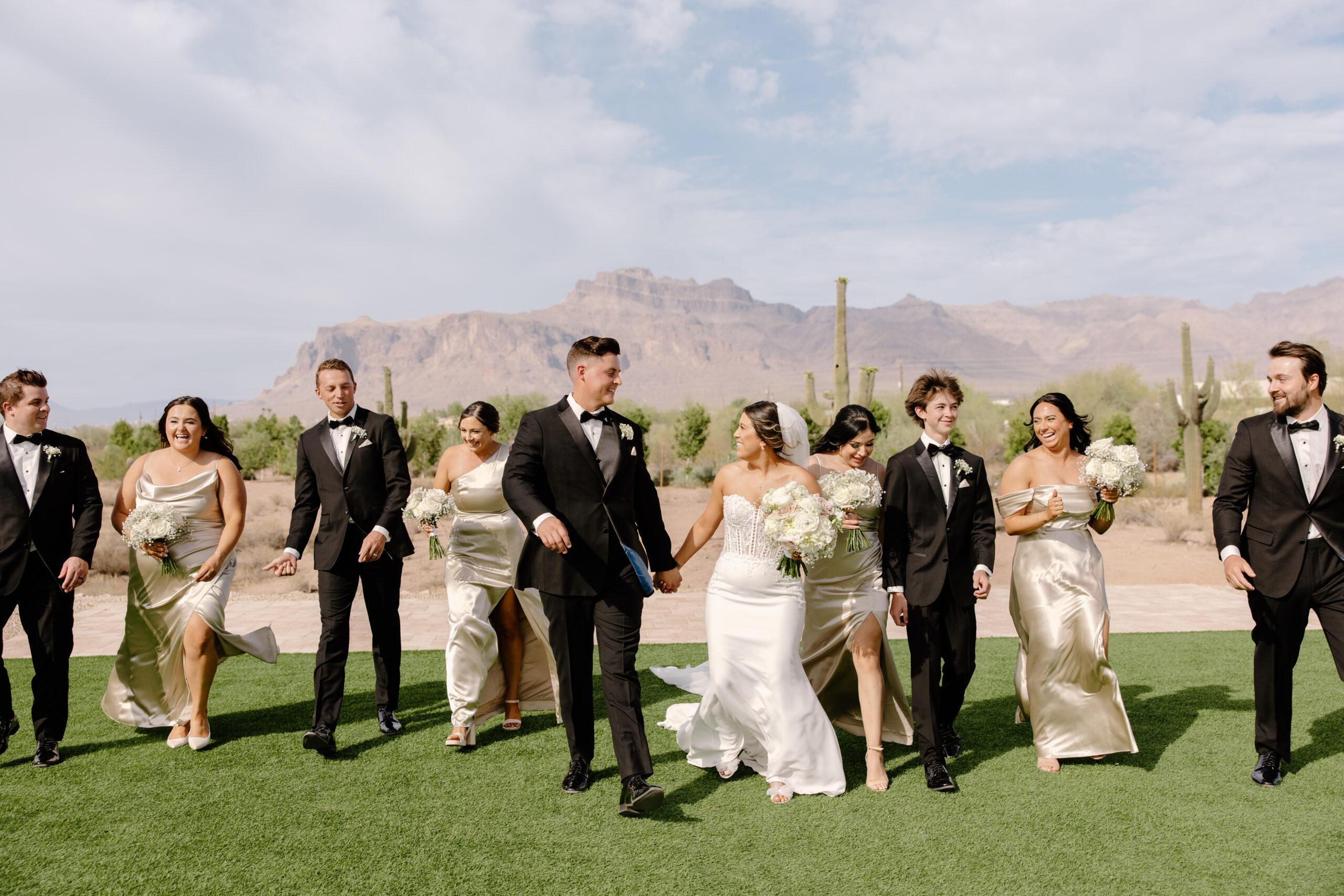 Bride and groom walking with their wedding party across green lawn with desert mountains and saguaros in the background during their Desert View wedding.