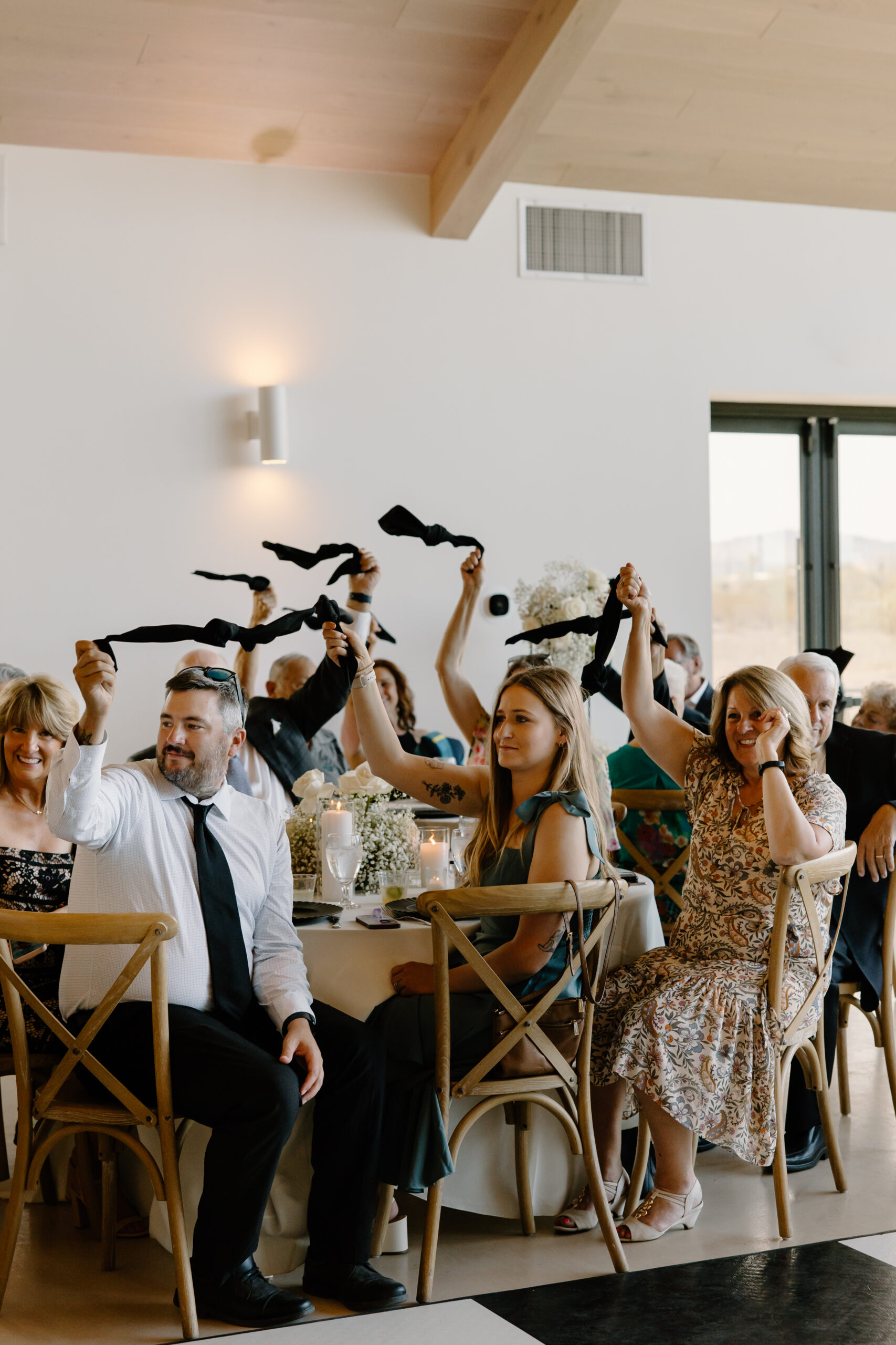 Wedding guests seated at reception tables raising black napkins in the air during a celebratory moment.