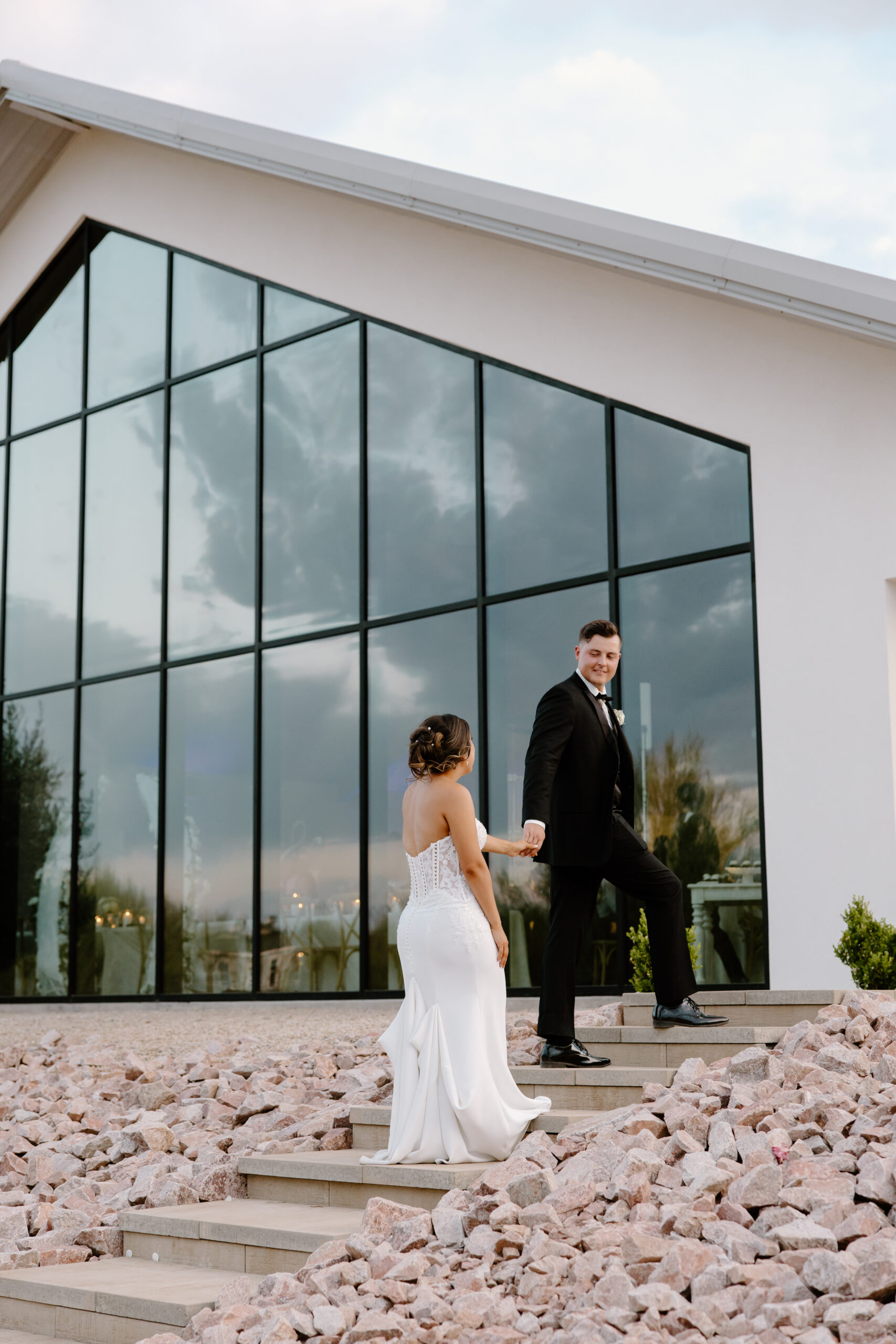 Bride and groom walking up modern stone steps outside the chapel at their Desert View wedding, with the tall glass windows reflecting the sky behind them.