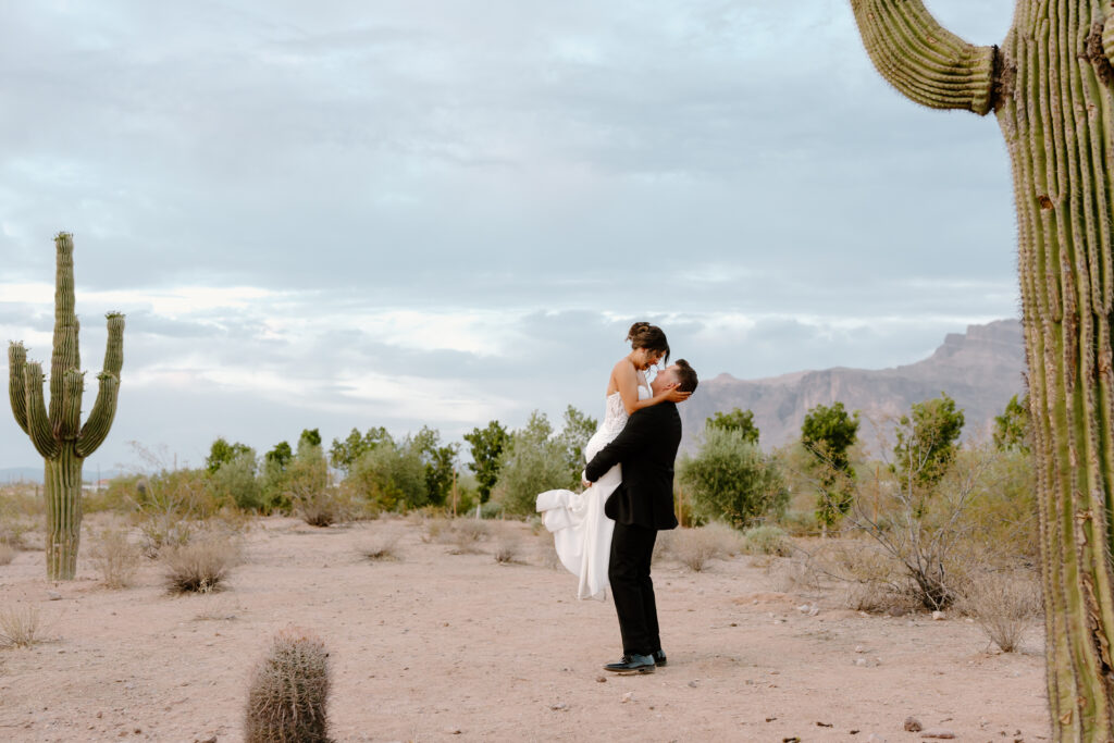Groom lifting the bride for a kiss in the Arizona desert surrounded by saguaros and mountain views after their wedding ceremony.